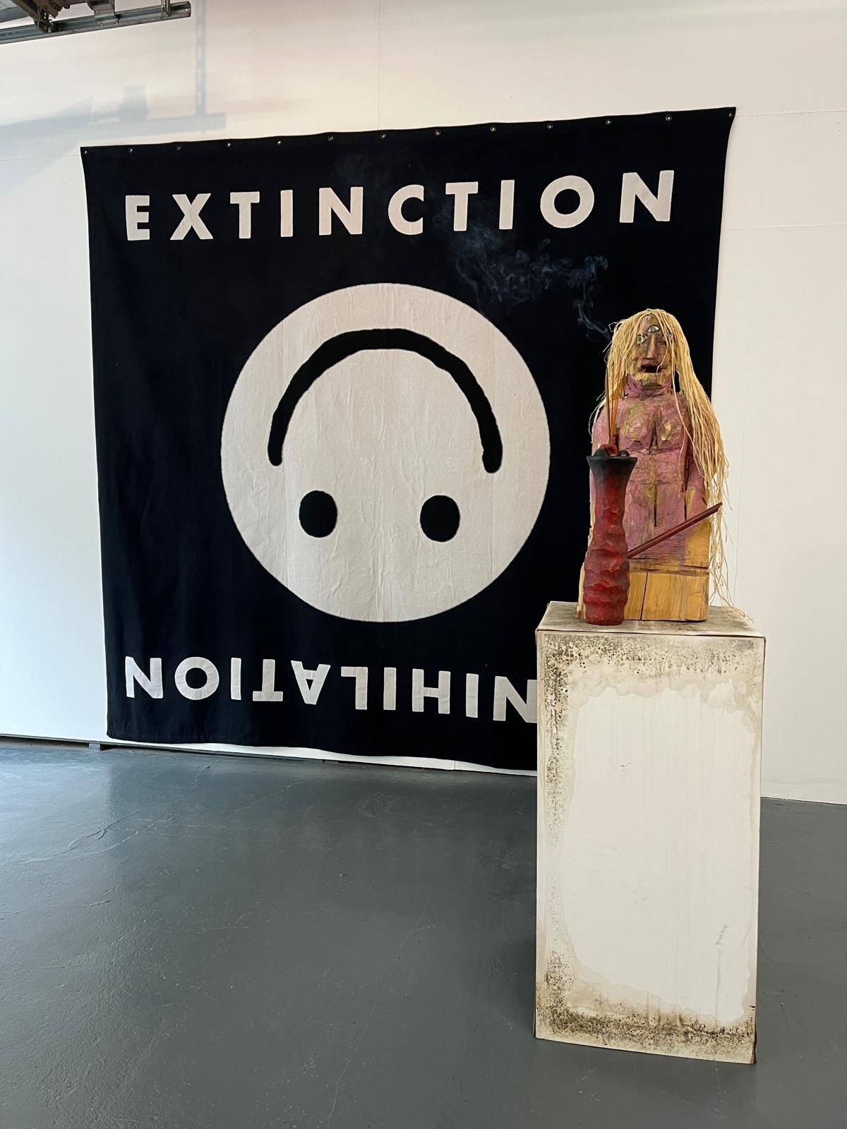 An art installation featuring a sculpture of a figure with long blonde hair and a pink dress, sitting on a wooden platform with a red vessel and a stick. Behind the sculpture is a black fabric banner with a white face symbol and the words 'Extinction