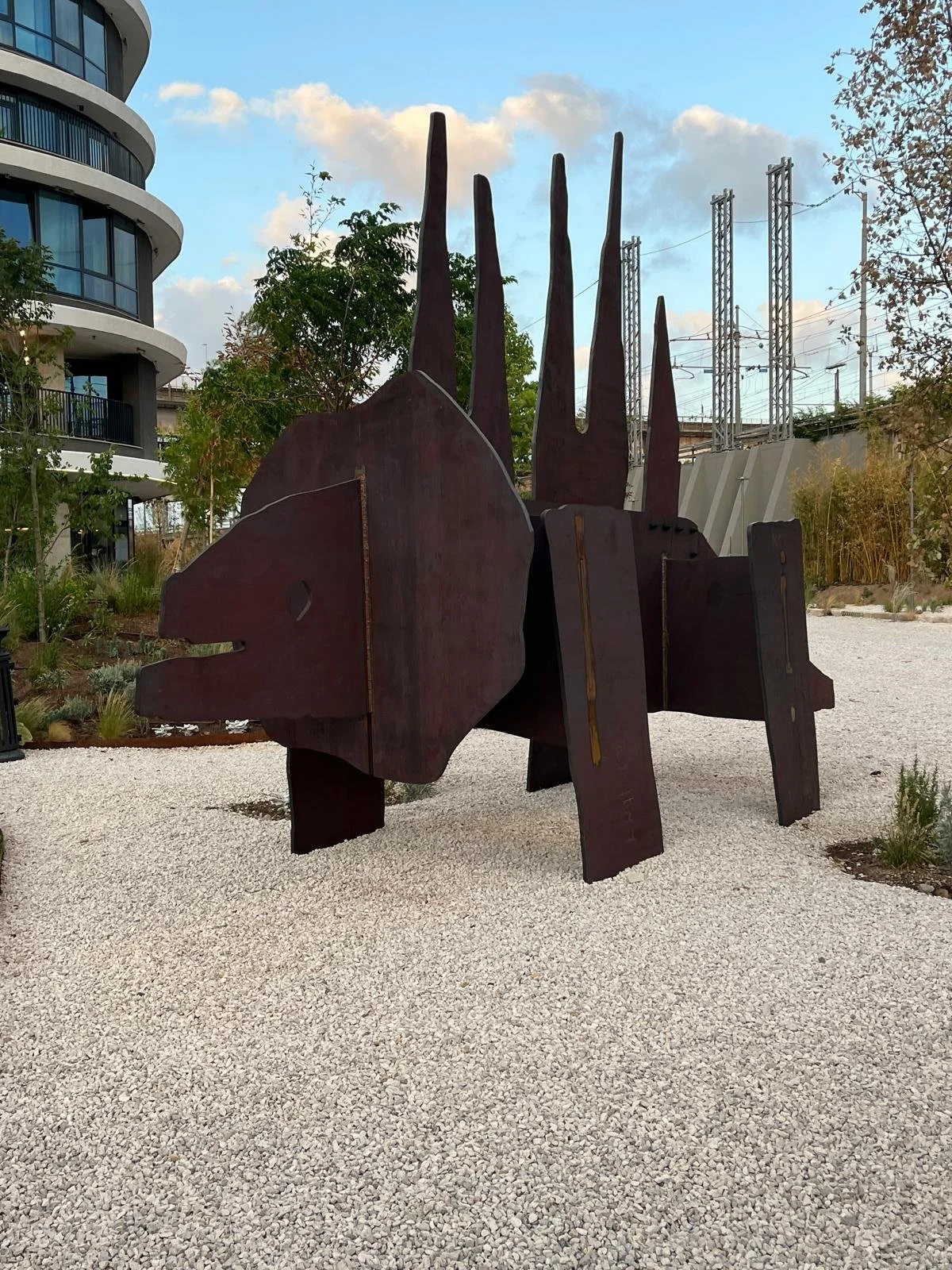 Abstract metal sculpture resembling a dog with pointed ears, made of large rust-colored metal panels, positioned on a gravel surface outdoors with modern building and power lines in the background.