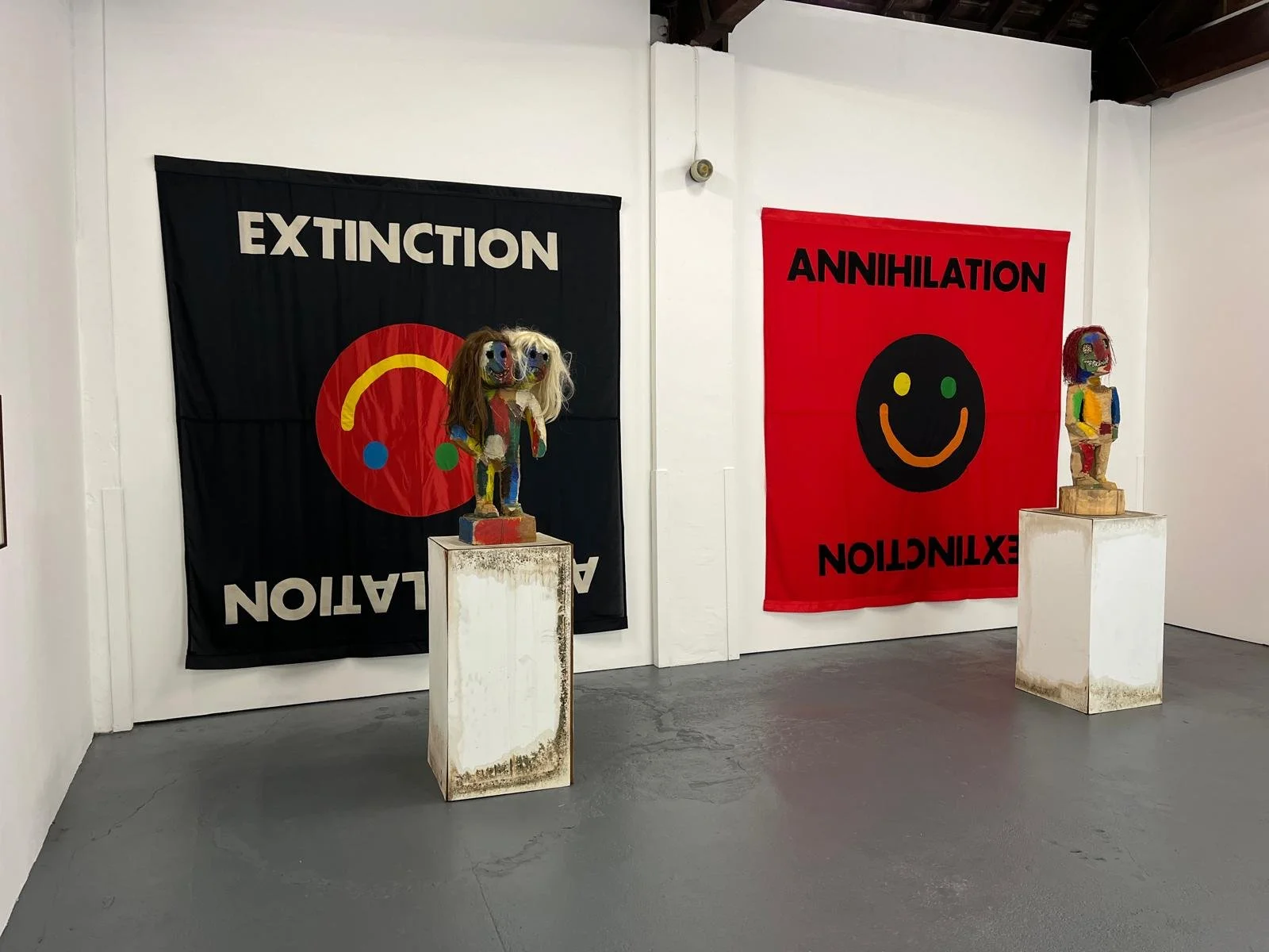 Art installation with two colorful face masks on sculptures, and two banners with black and red backgrounds displaying words "Extinction" and "Annihilation" along with smiley face symbols.