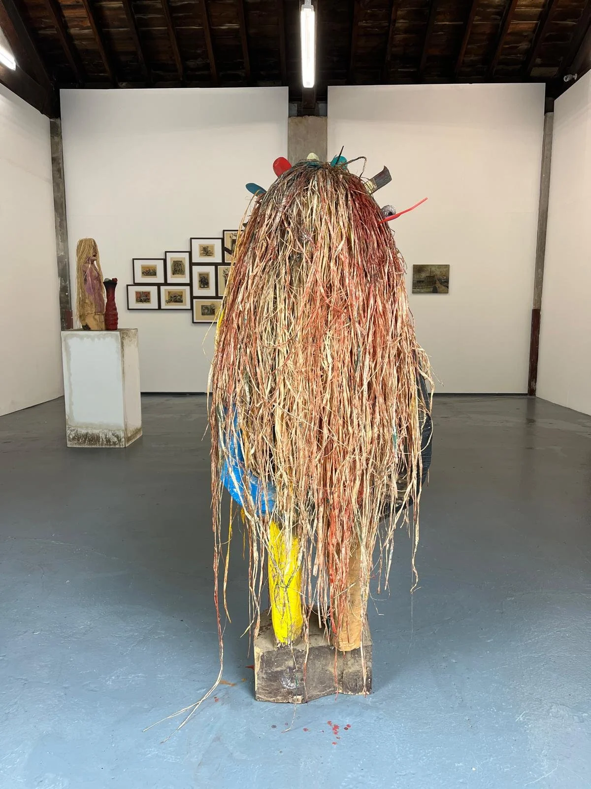 An art installation featuring a figure covered with long, straw-like materials and colorful objects protruding from the top, displayed in an art gallery with framed artworks on the walls.