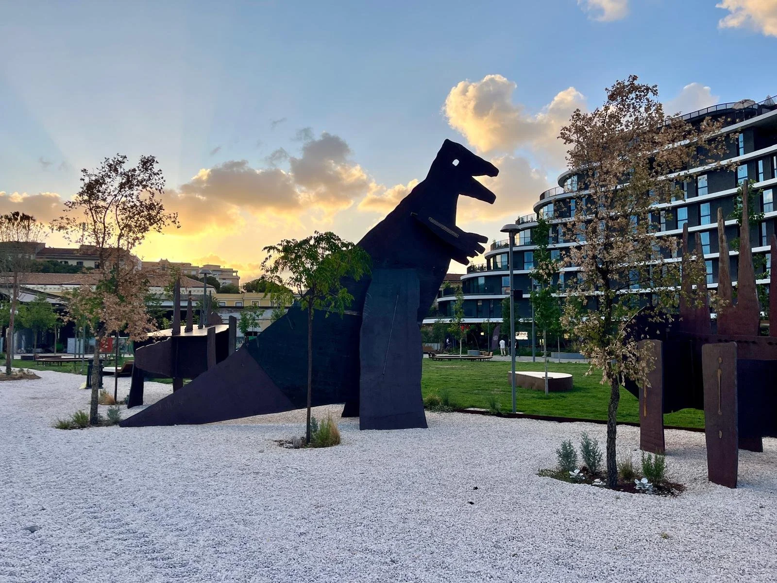 Large black abstract sculpture resembling a dragon or sea serpent in a landscaped park with trees, modern apartment buildings, and a sunset sky in the background.