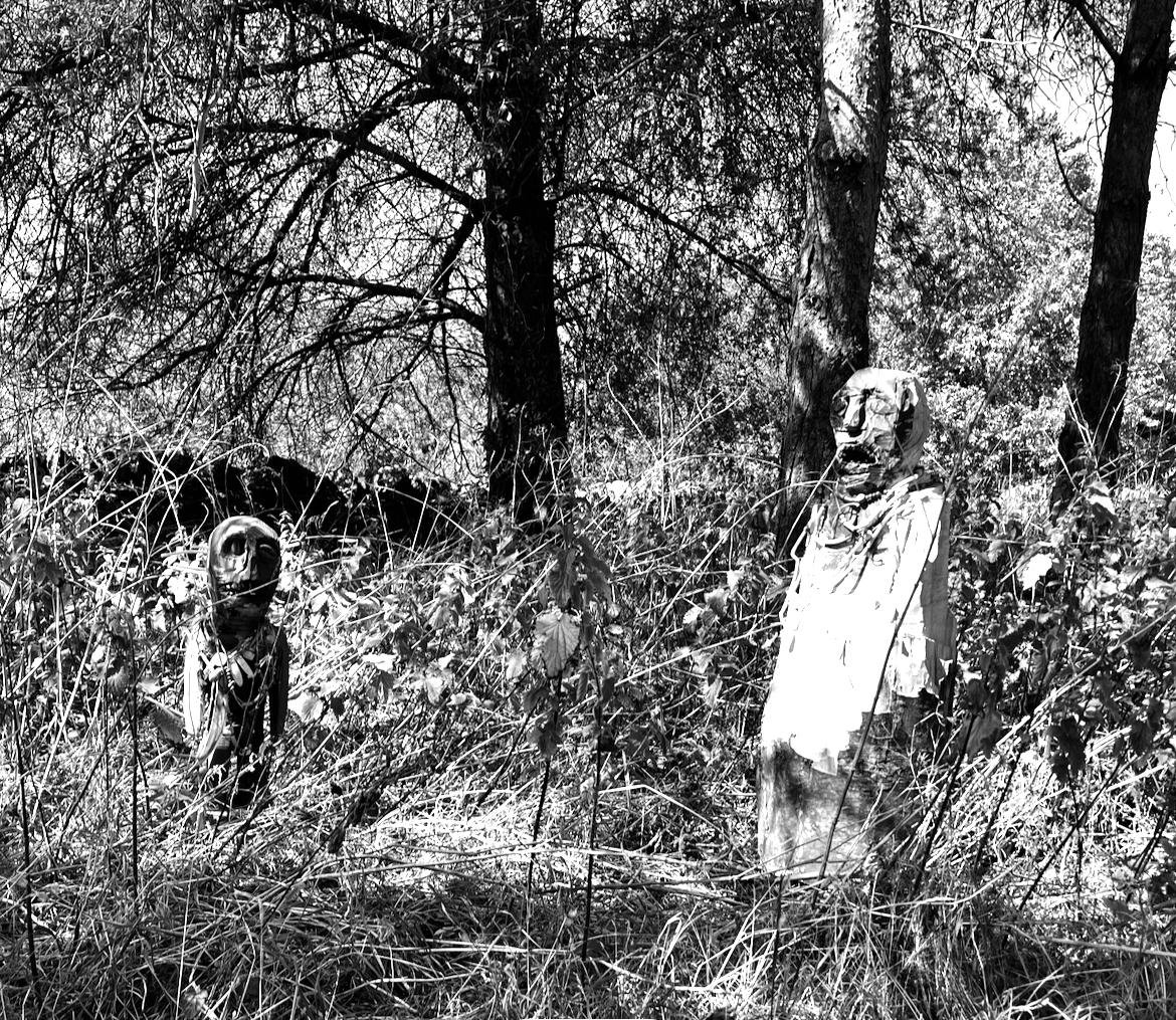 Black and white photo of two animal sculptures made from tree trunks and branches, set in a wooded area with tall grass and trees.