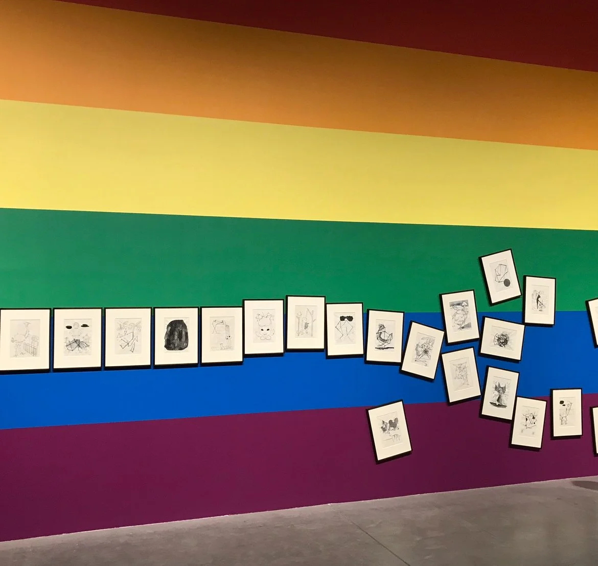 Wall with rainbow colors background displaying framed black and white abstract sketches and drawings, some tilted or scattered.