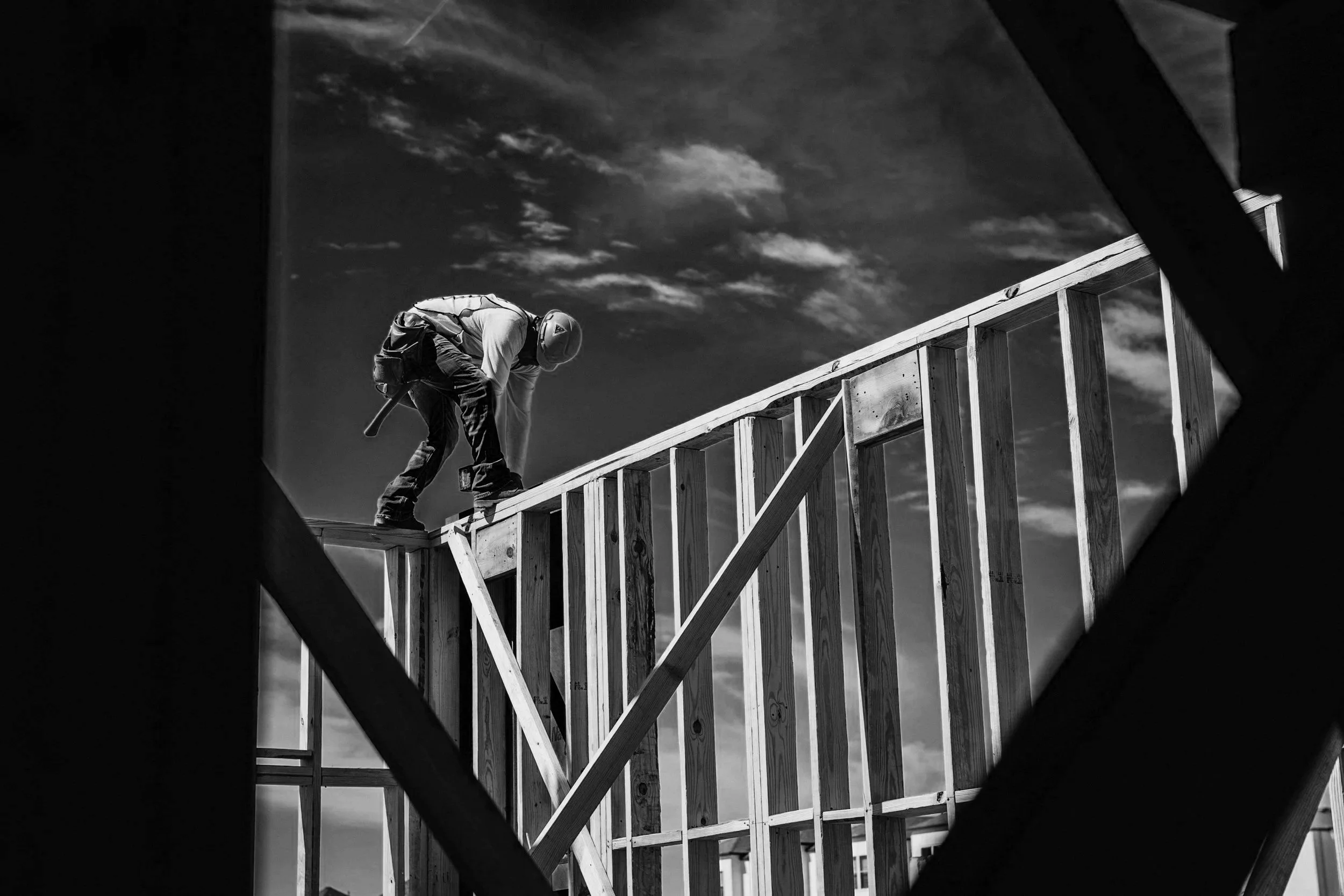 A construction worker wearing a helmet and safety harness working on the wooden framing of a building under a partly cloudy sky.