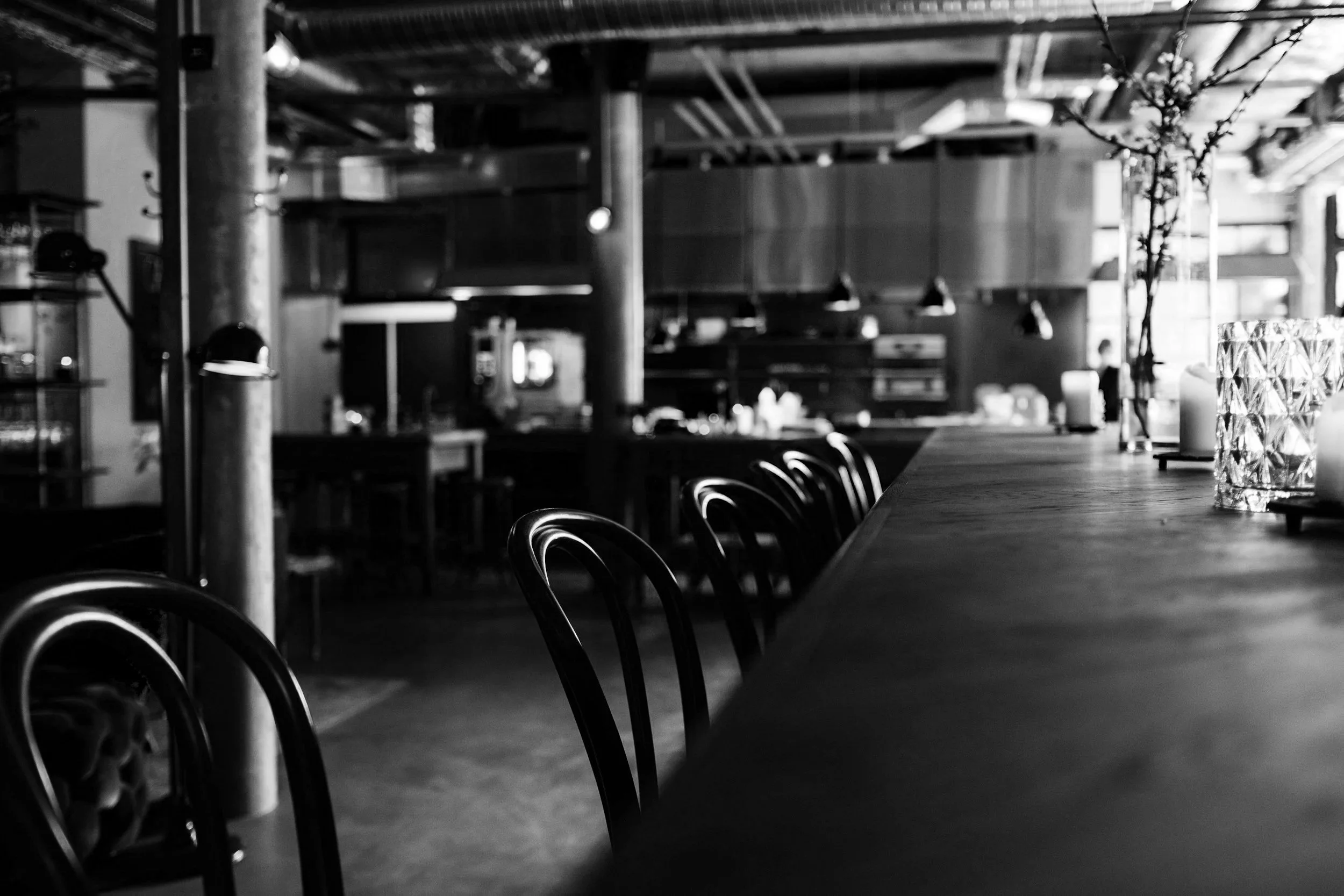 Empty restaurant with chairs lined up along a long wooden table, candles and flowers on the table, industrial-style interior with exposed pipes and beams, black and white photo.