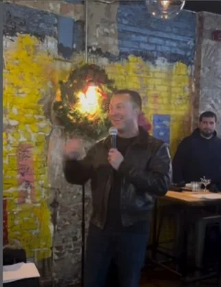 Man speaking into a microphone at a decorated indoor event with a yellow and brick wall background.