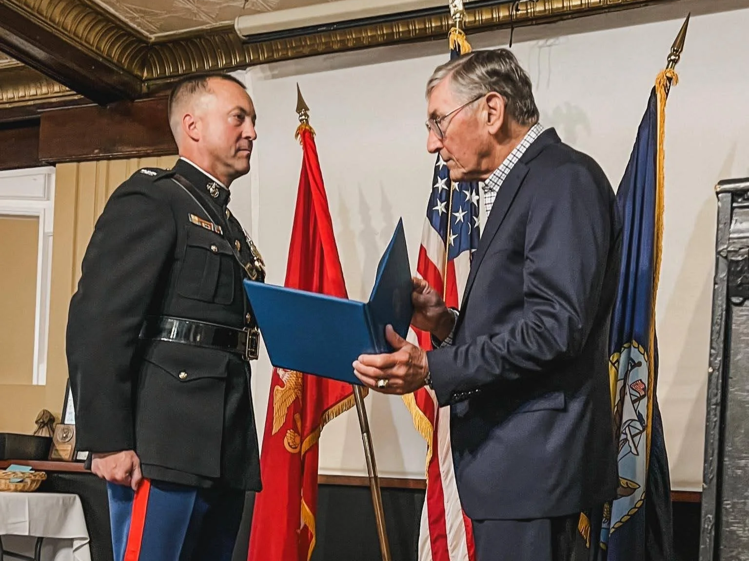 A uniformed military officer receiving a medal or award from an older man in a suit, with American and military flags in the background, in an indoor setting.