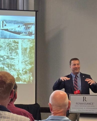 A man in a suit giving a presentation in a conference room, with a projection of an airplane on the screen behind him and an audience listening.