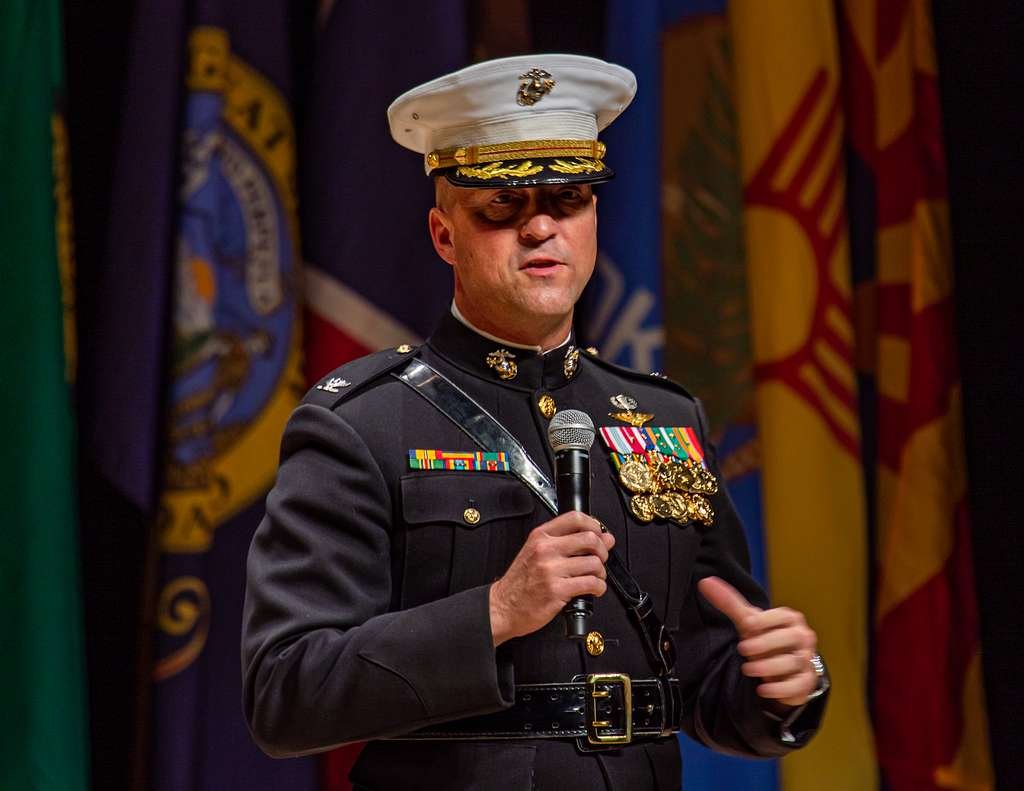 A man in a United States Marine Corps dress uniform speaking into a microphone with military flags in the background.