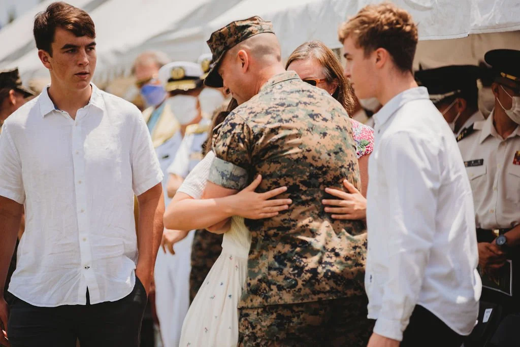 Military personnel and civilians embrace and console each other at an outdoor event, with others wearing masks in the background.