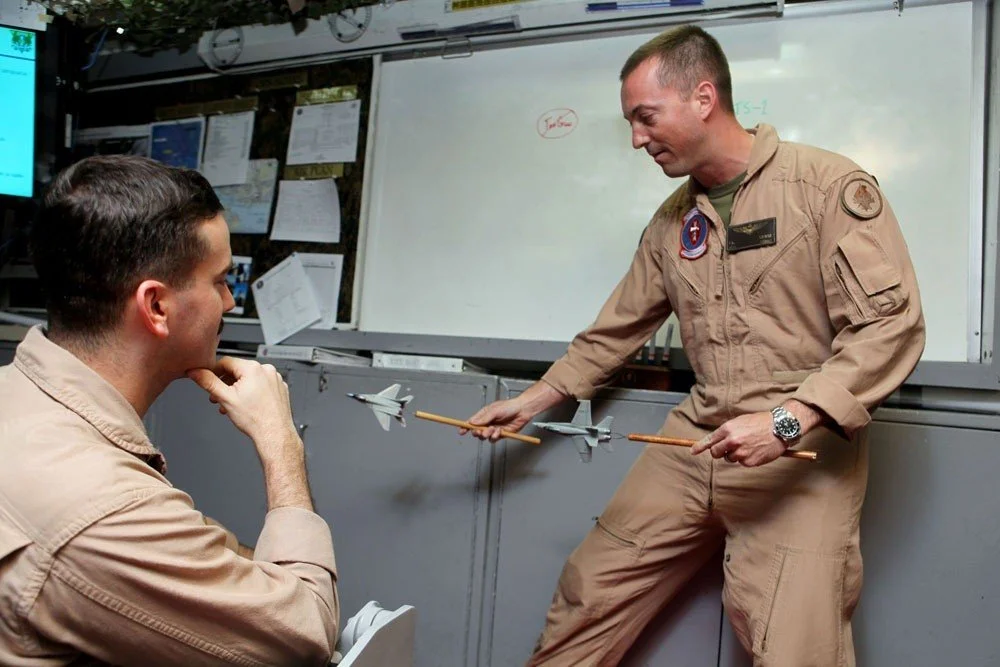 Two men in military uniforms in a briefing room, one demonstrating a model of an aircraft model to the other.