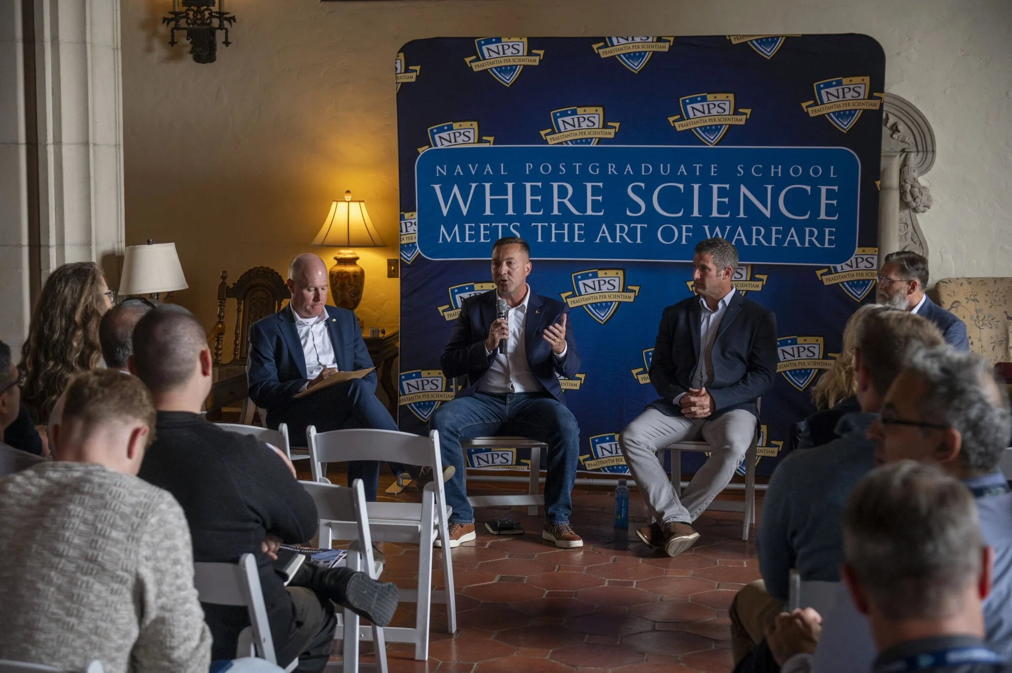 Panel discussion at Naval Postgraduate School with five men seated on stage in front of a blue backdrop. One man is speaking into a microphone, others are listening. Audience seated in white chairs facing the stage. Warm lighting, vintage lamps, and 