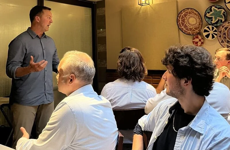 A man giving a presentation or speech to a group of people seated at a table in a restaurant or conference room. The audience appears engaged, with some looking at the speaker and others looking away. There are decorative plates on the wall behind th