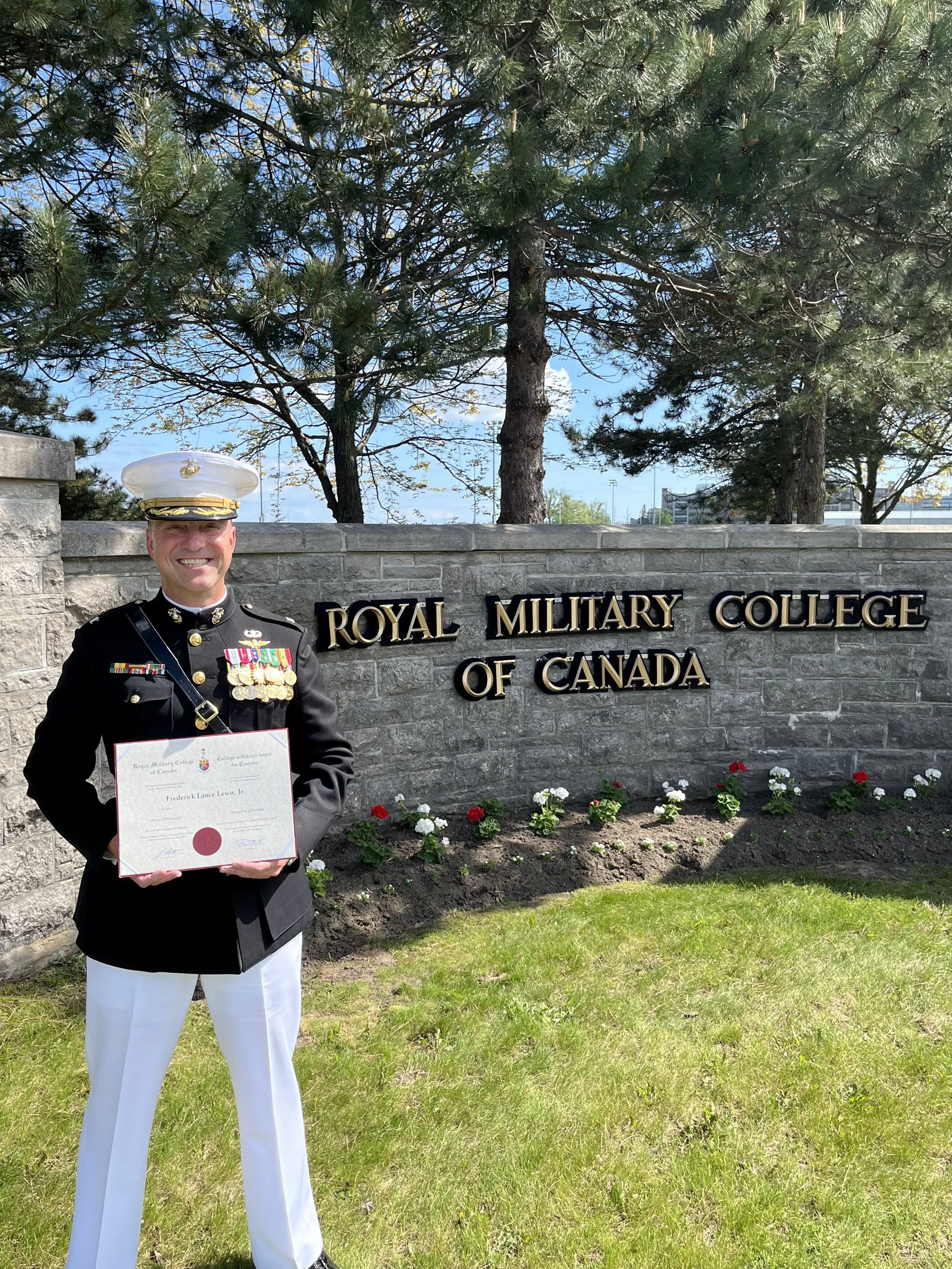 A person in a military uniform holding a certificate standing in front of a sign that reads 'Royal Military College of Canada' with trees and a garden in the background.
