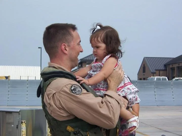 A man in military uniform holding a young girl in a floral dress and smiling at her outdoors.