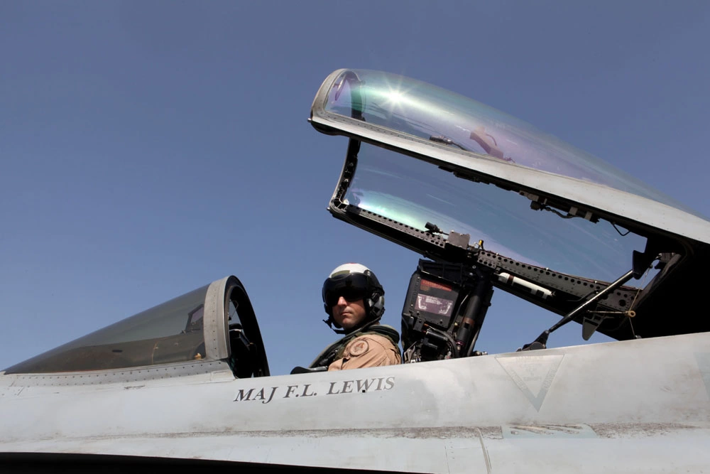 Military pilot wearing a helmet seated inside the cockpit of a fighter jet with the canopy raised.