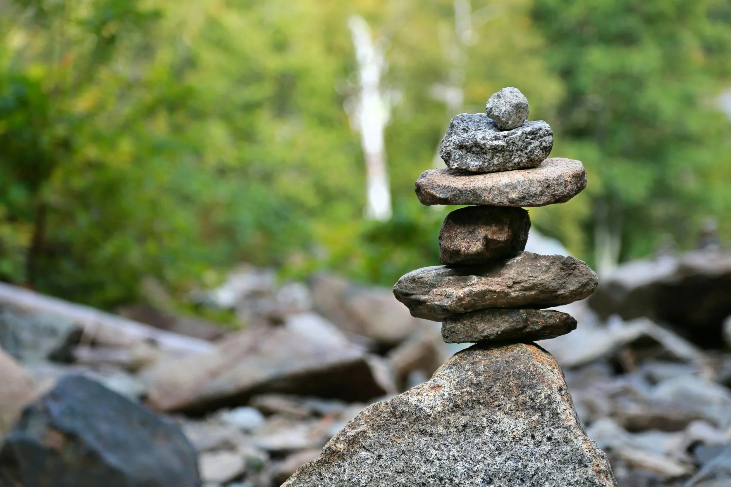 A stack of seven rocks balanced on top of each other in a natural outdoor setting with green foliage in the background.