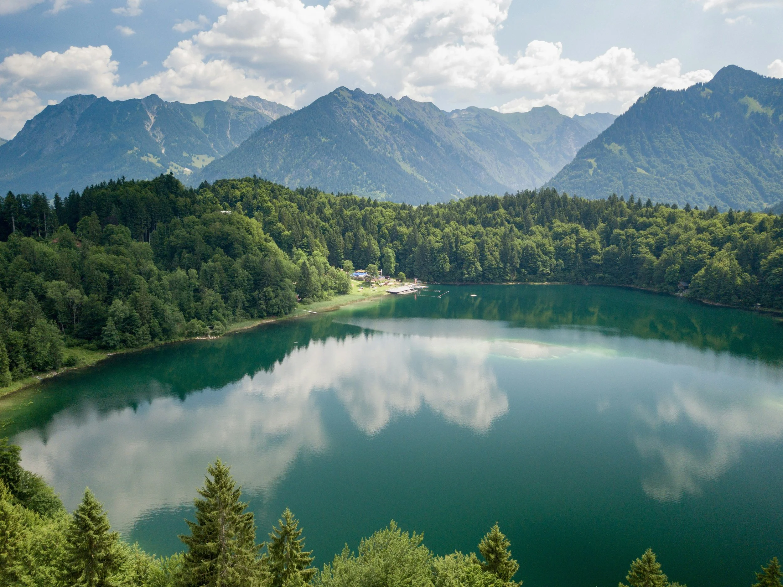 A scenic view of a mountain lake surrounded by dense green forest, with mountains in the background under a partly cloudy sky.