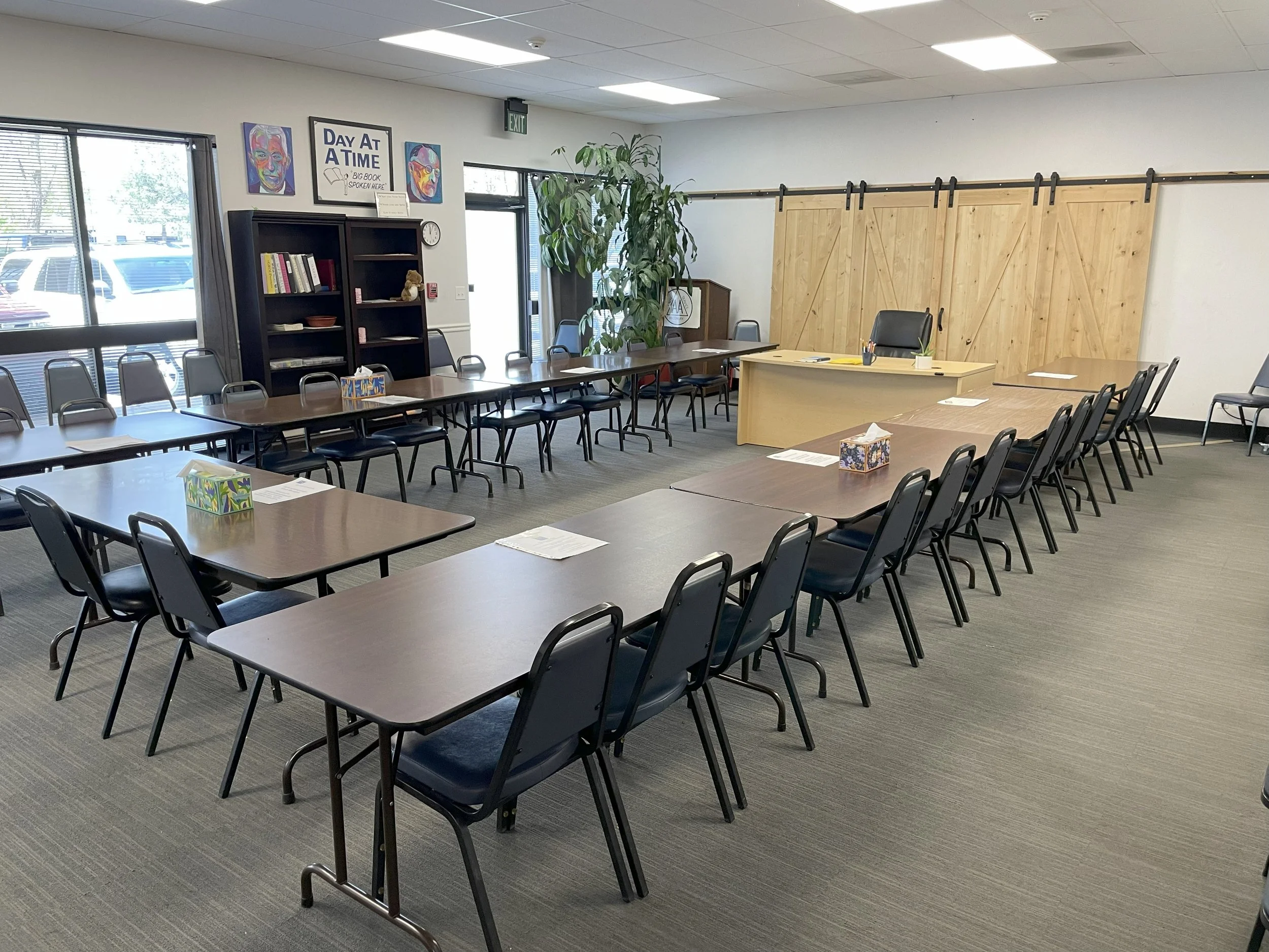 An empty conference room with several rectangular tables arranged in a U-shape, surrounded by black chairs. There are tissue boxes and papers on the tables. The room has large windows, a bookshelf, a large plant, and a wooden sliding barn door at the back. There are colorful paintings and a sign on the wall.