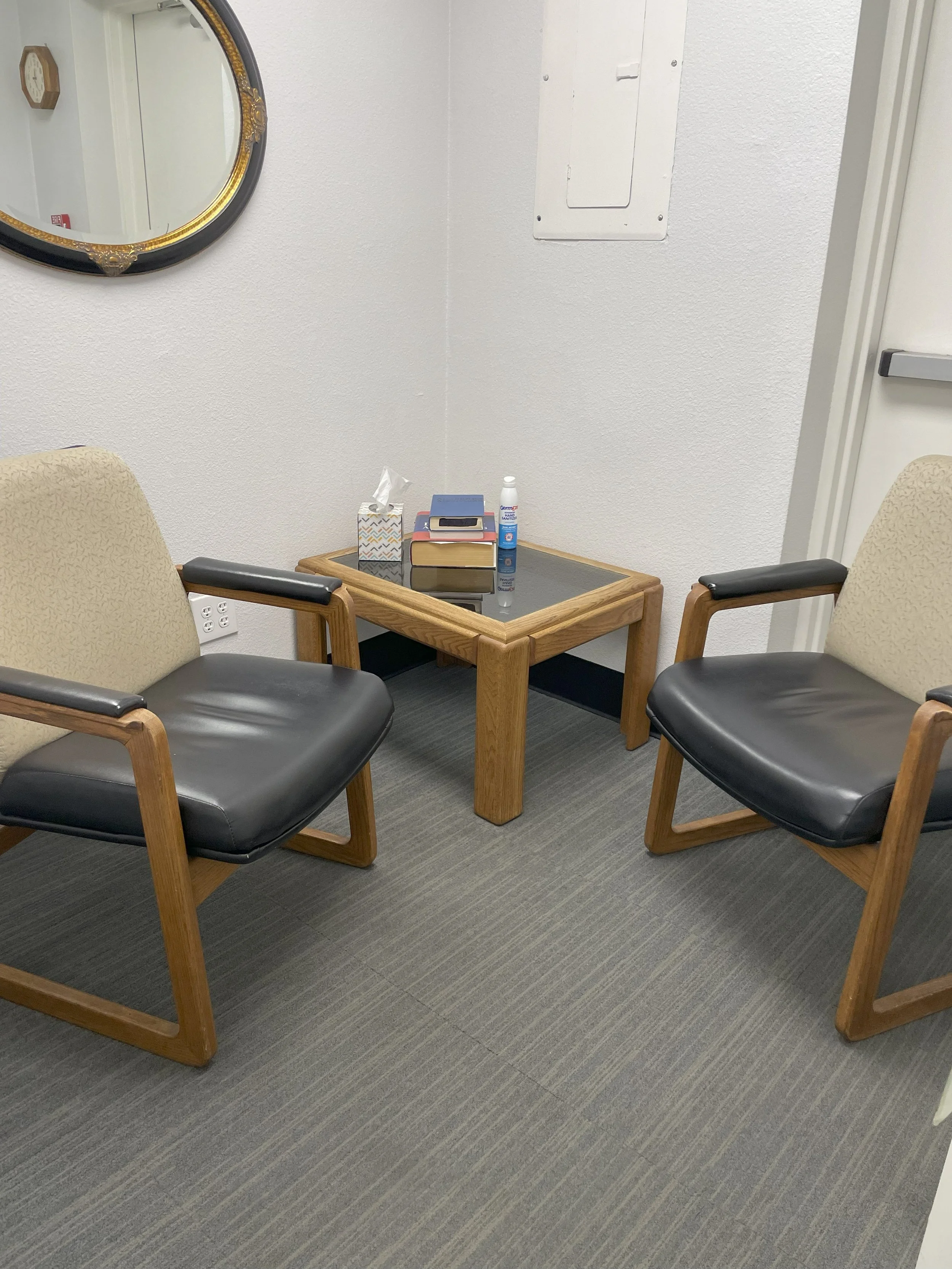 Two beige and black chairs with wooden frames and padded armrests facing each other, with a small wooden table in between. The table has tissues, a box, a small bottle, and some books or notebooks on it. There is a mirror on the wall behind the chairs and a white electrical panel on the wall, with a white door to the right.