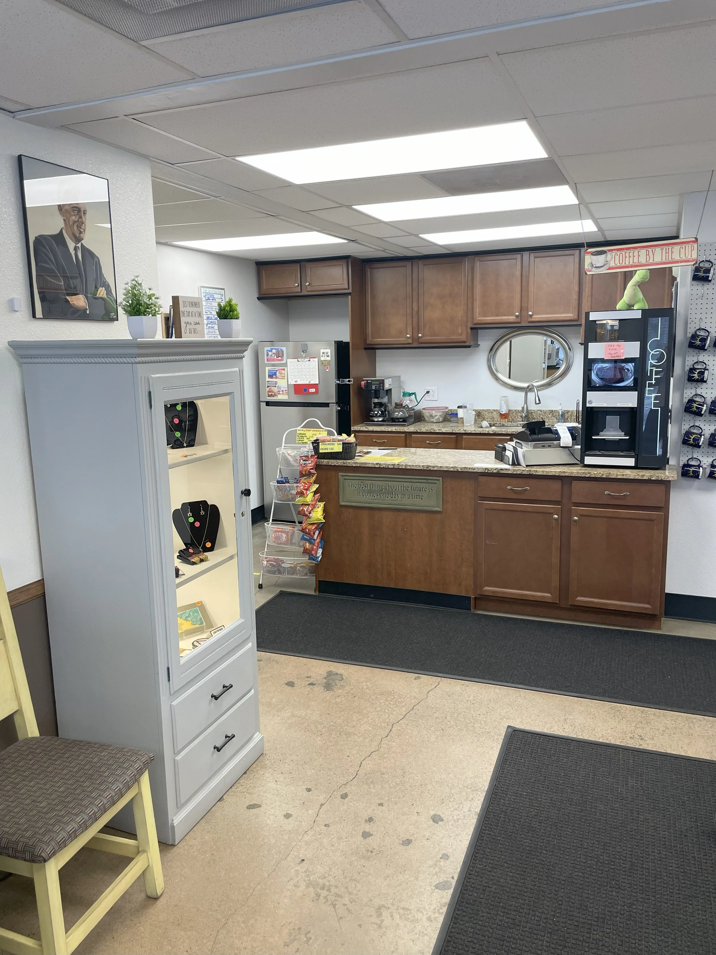 Coffee shop kitchen area with wooden cabinets, coffee machine, microwave, refrigerator, and decorative wall art.