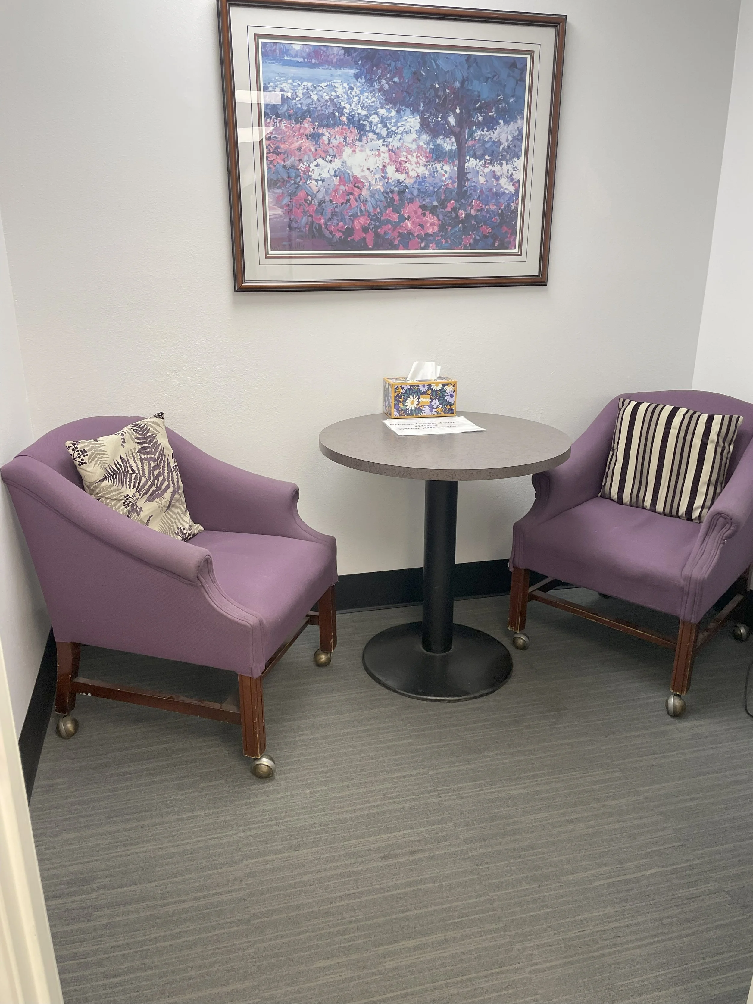 A small round table with two purple armchairs on either side, each with a decorative striped or leaf-patterned pillow. On the table, there's a box of tissues and a paper. Above, a colorful framed landscape painting of flowers and trees is hung on the wall.