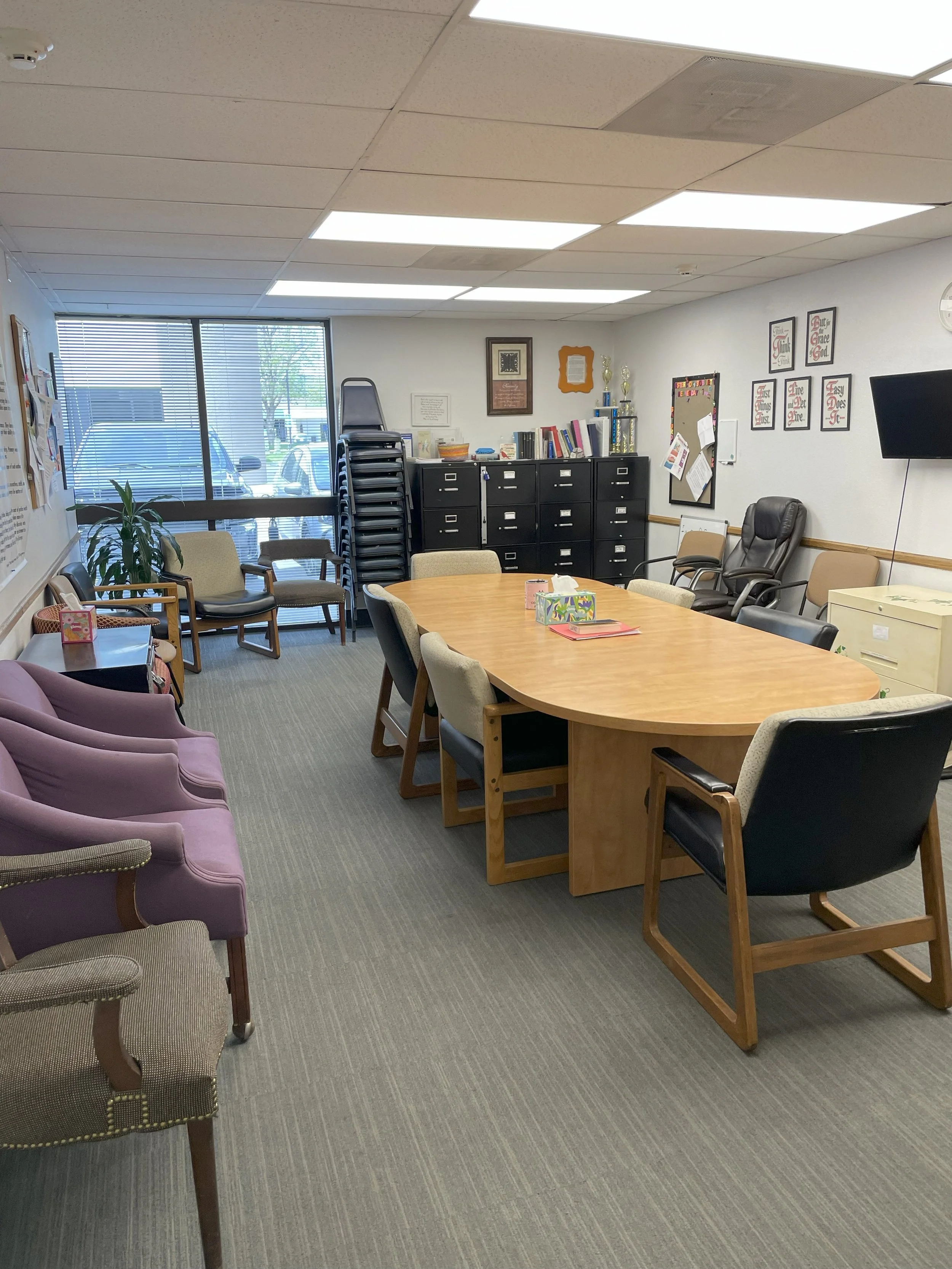 An office meeting room with a large wooden table surrounded by assorted chairs, a black filing cabinet, tiered chairs stacked near window, purple and gray armchairs, and various wall decorations.