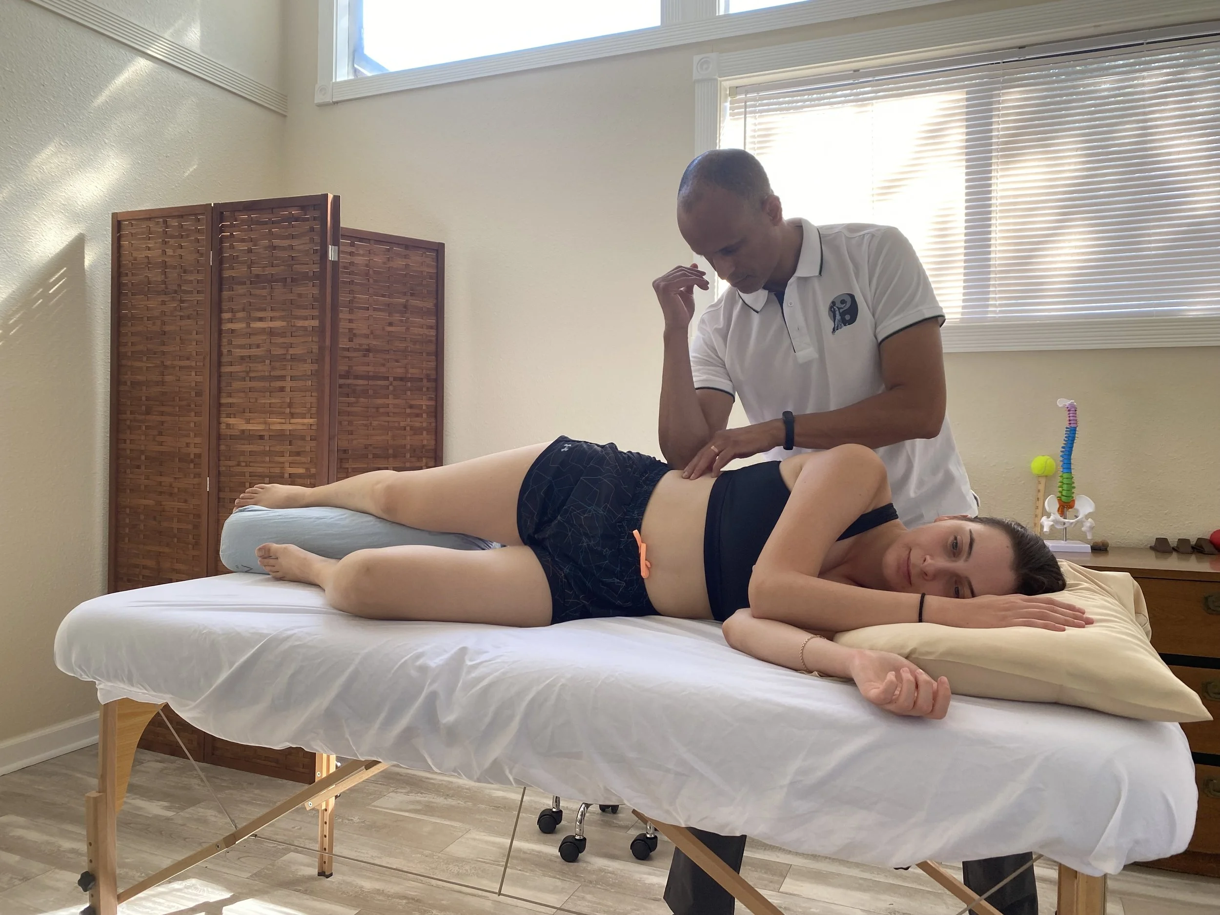 A person receiving back massage therapy from a therapist in a bright room with large windows, a wooden room divider, and massage equipment.