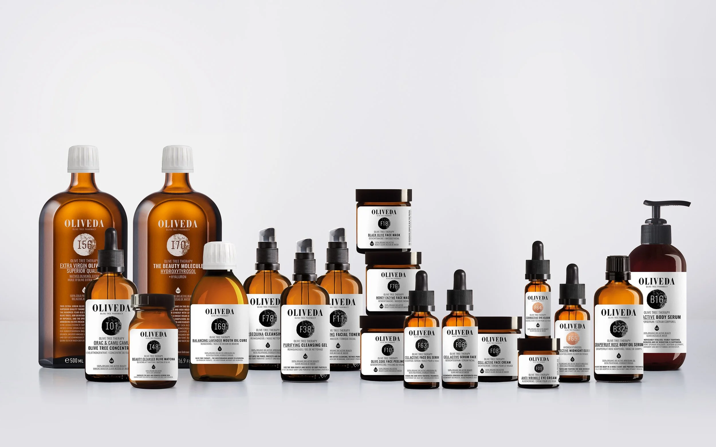 A collection of skincare products from Oliveda, including bottles, jars, and dropper bottles, arranged on a white surface against a plain white background.