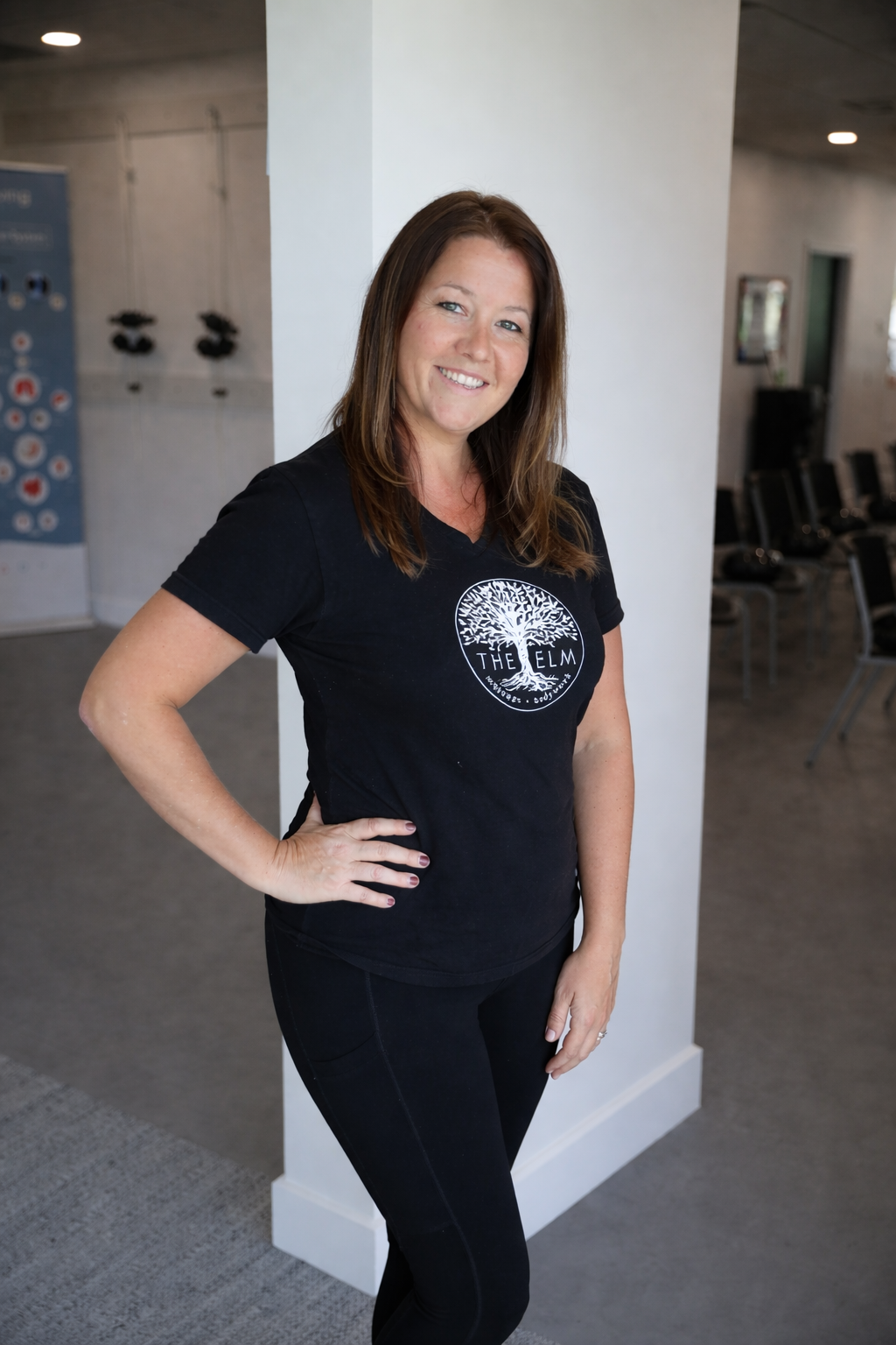 A woman with brown hair and a black T-shirt that has a tree logo and the words "The Elm" standing in a room with chairs, facing the camera, smiling, with her hand on her hip.