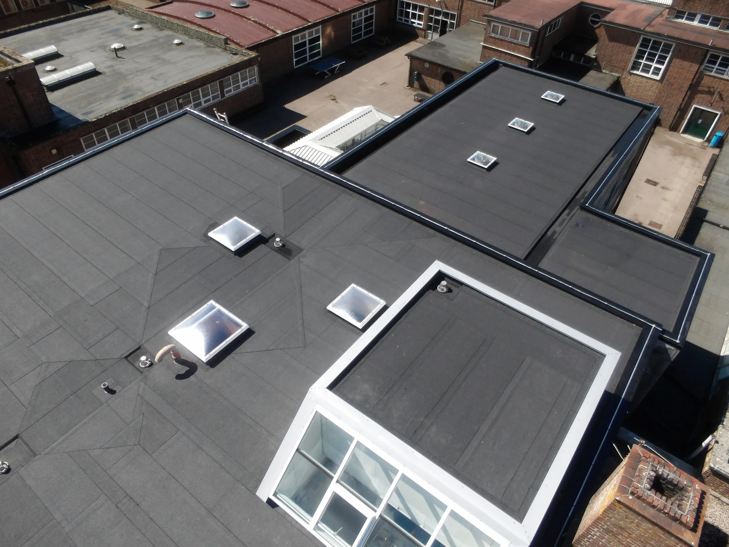 Aerial view of flat rooftops with vents, skylights, and a section with white framing, in a city setting.