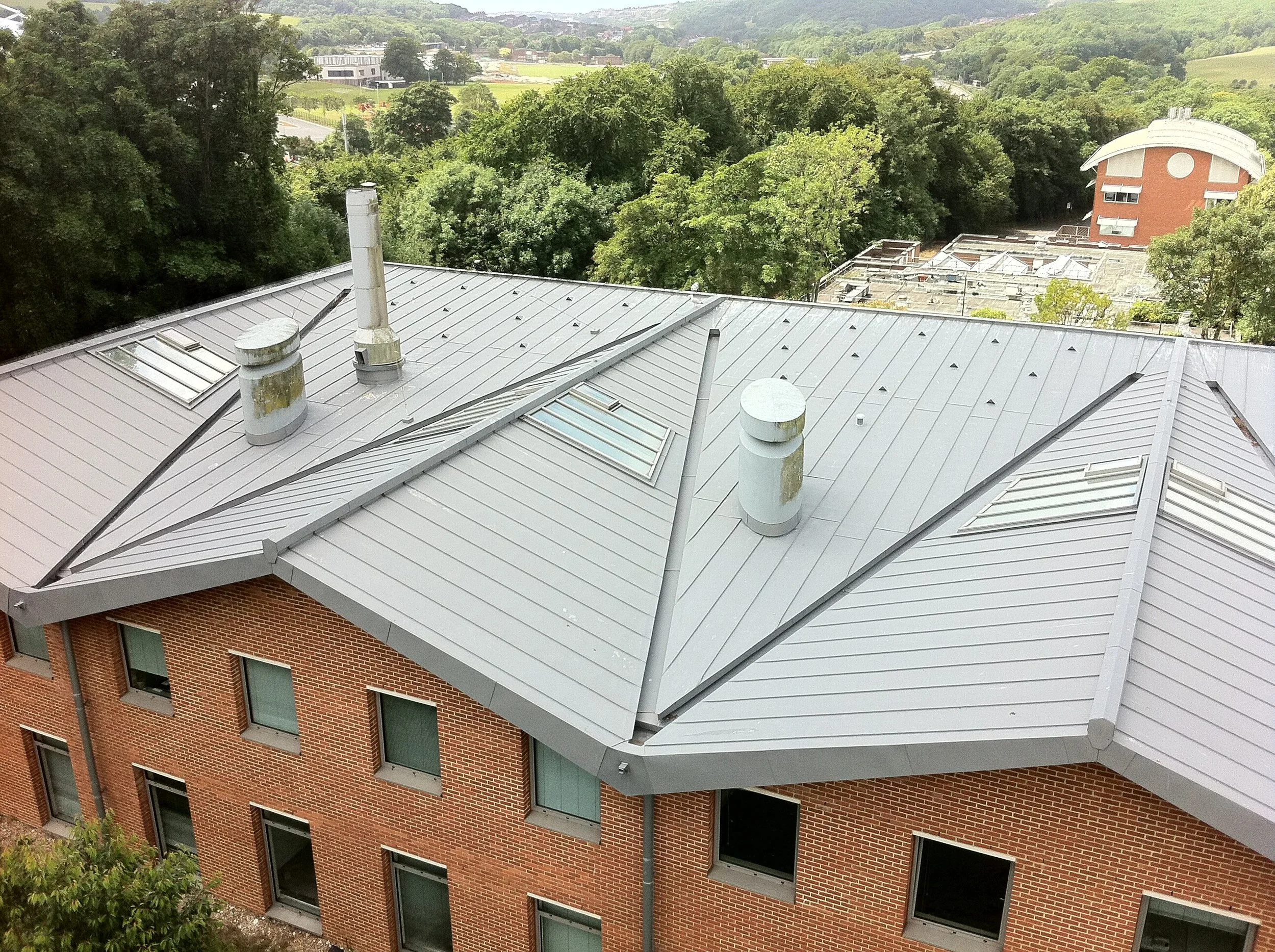 Aerial view of a building's gray metal roof with multiple vents and skylights, surrounded by green trees and some distant buildings.