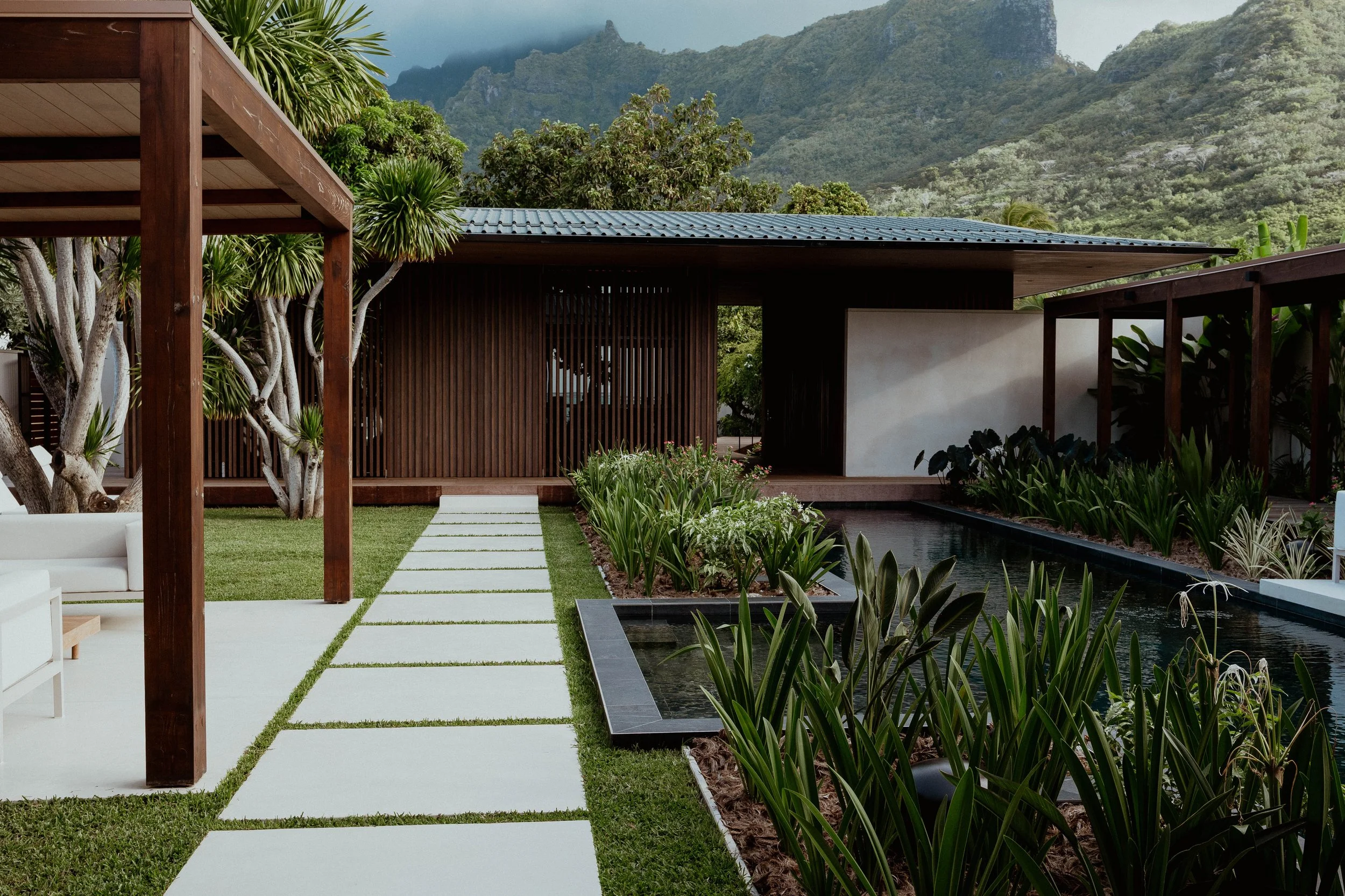 Un jardin moderne avec une allée de dalles blanches, une piscine et une maison en bois avec un toit en métal, entouré de végétation luxuriante et de montagnes en arrière-plan.