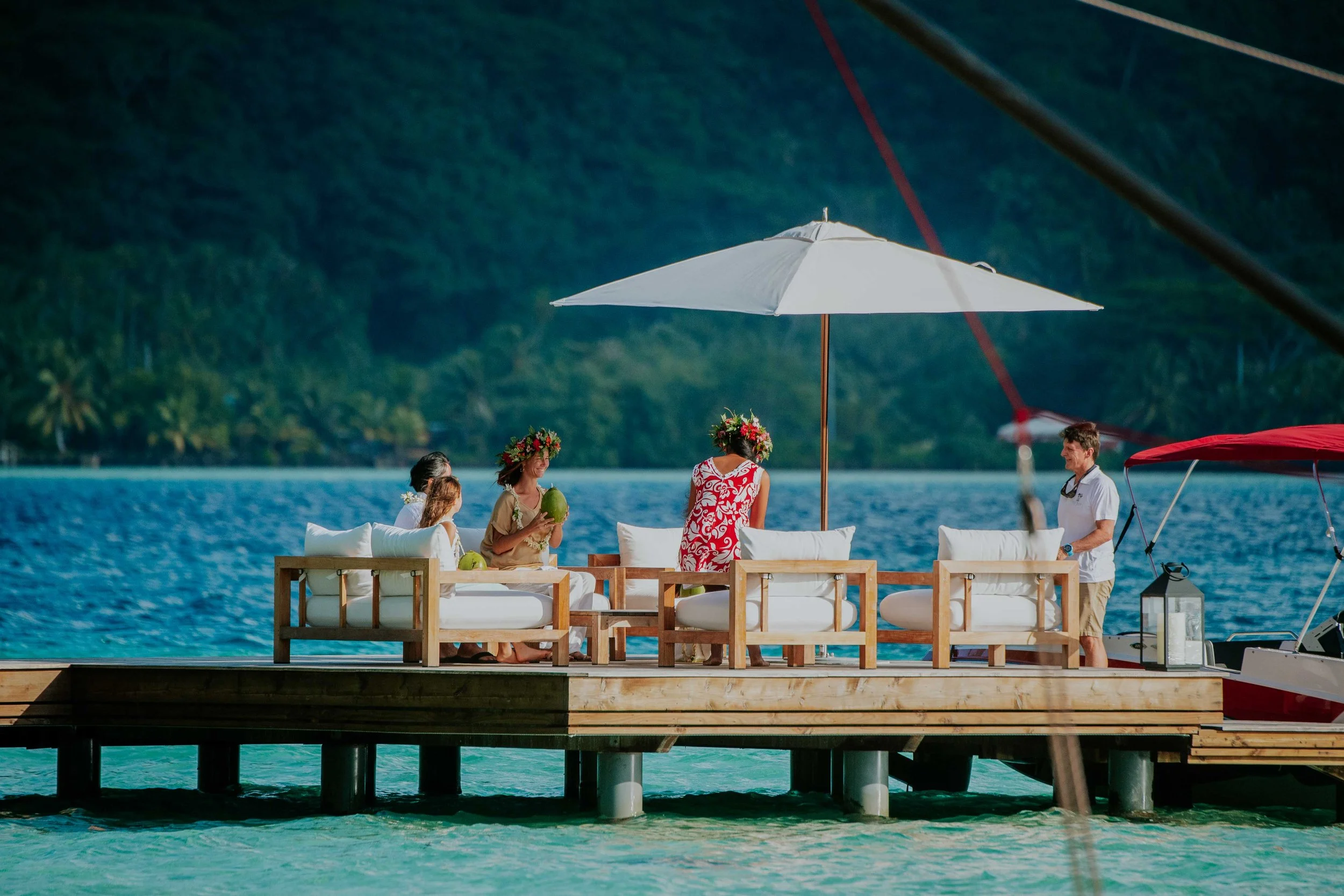 Groupe de personnes sur une terrasse en bois au bord de l'eau, avec parasol blanc, cocotiers et montagnes en arrière-plan, ambiance détendue et tropicale.
