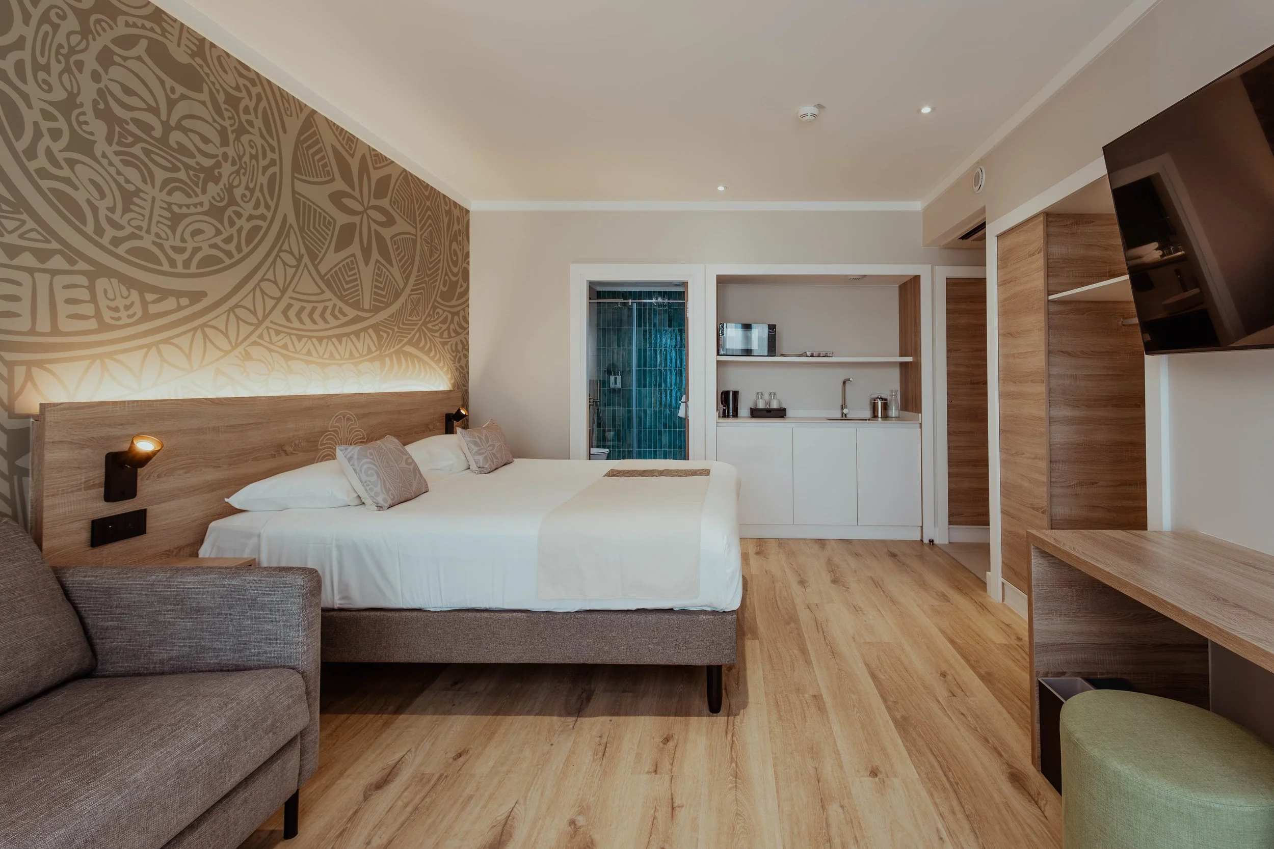 Chambre d'hôtel moderne avec lit double, sofa gris, bureau en bois, kitchenette, télévision suspendue, mur en bois, sol en bois, porte de la salle de bain en verre, lampe de lecture.
