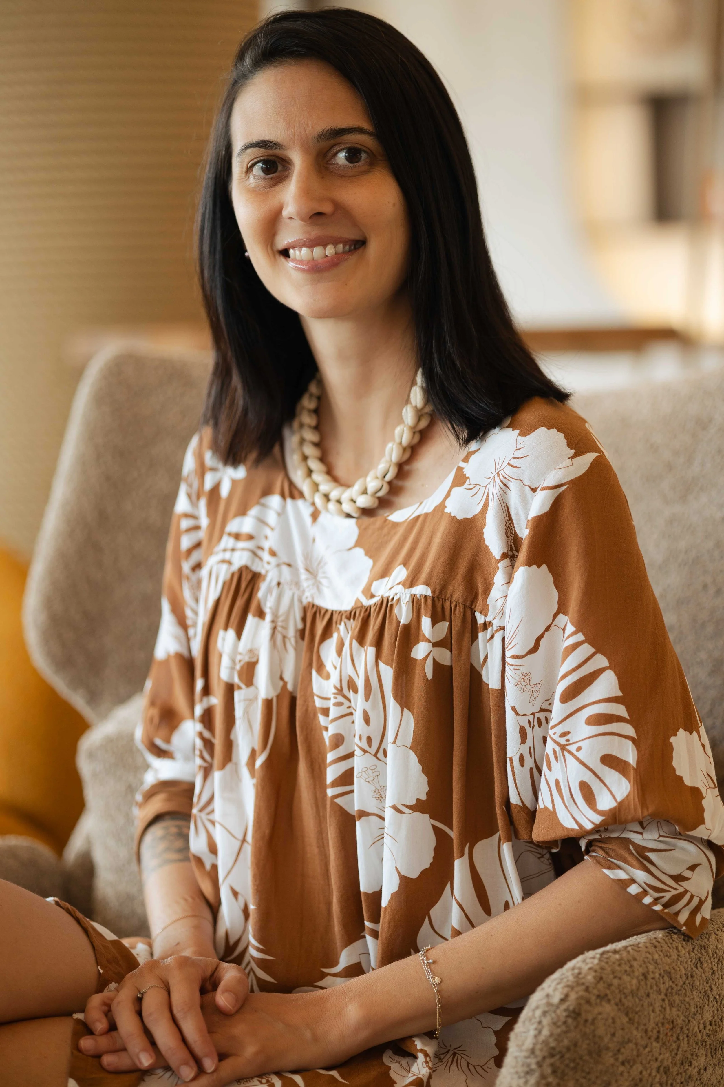 Une femme assise sur un canapé, souriante, portant un collier de perles blanches et une robe à motif floral marron et blanc.