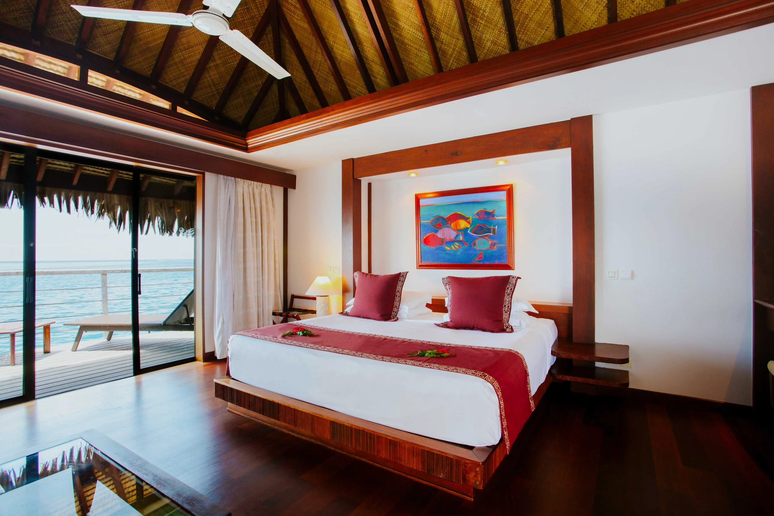 Chambre d'hôtel avec lit double, décoration en bois, vue sur la mer, porte-fenêtre avec balcon, peinture de poissons dans l'eau, linge de lit avec coussins rouges.