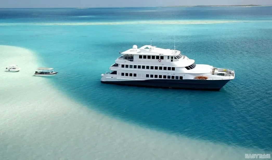 Un yacht blanc de grande taille navigue dans une mer turquoise, avec deux petits bateaux à proximité dans l'eau claire.