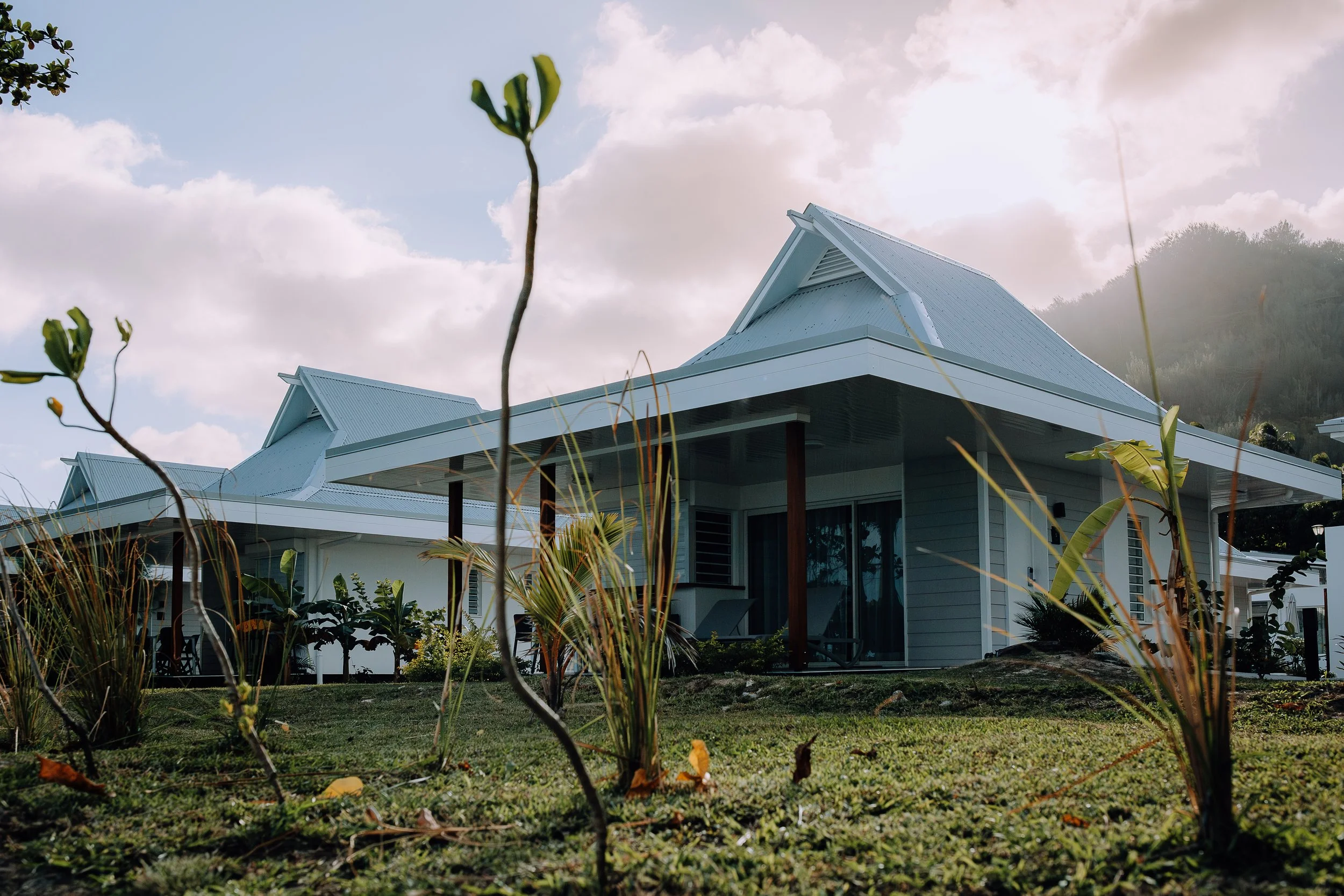 Une maison moderne avec un toit en pente, entourée de végétation, sous un ciel partiellement nuageux.