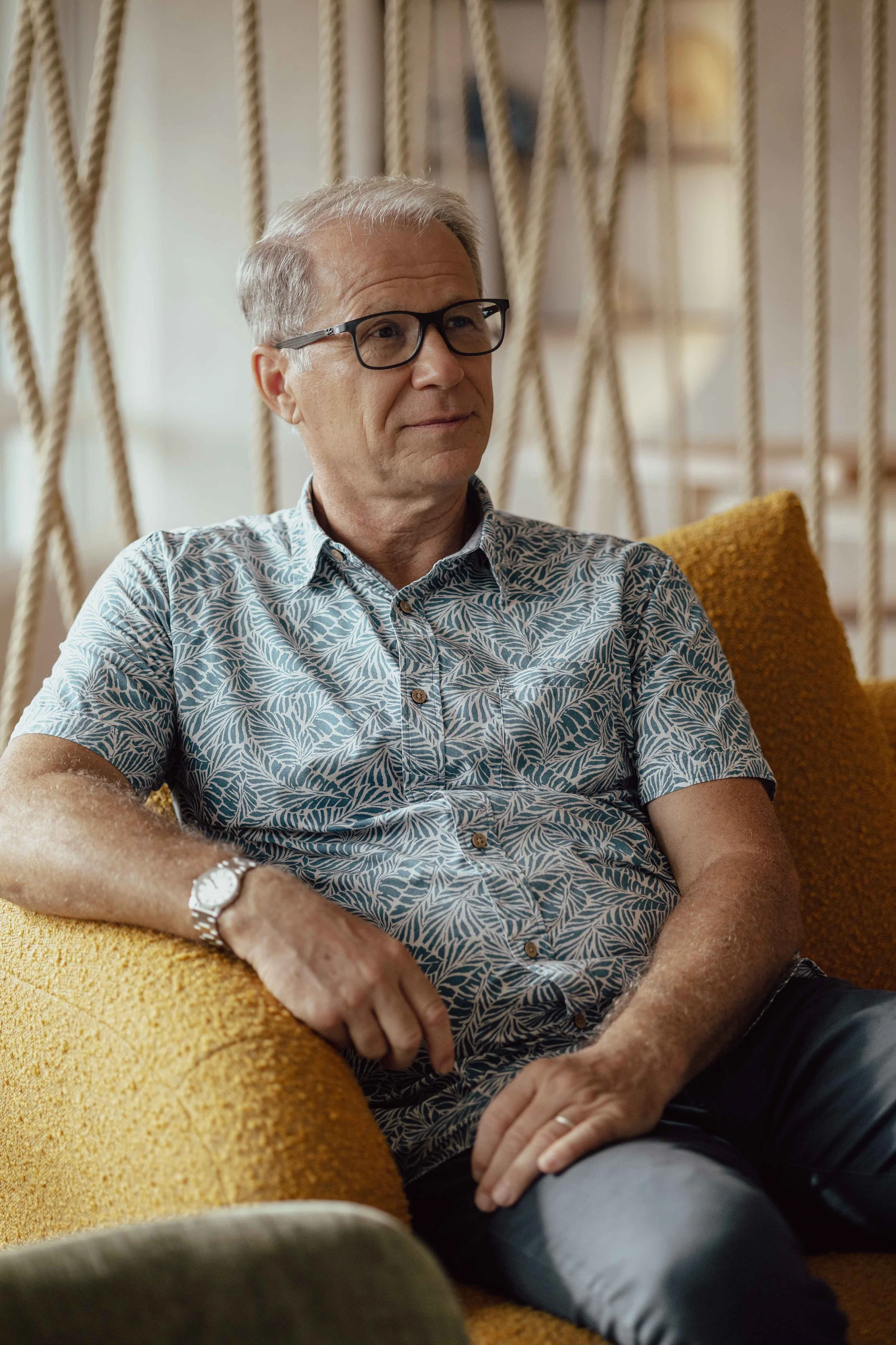 Un homme âgé avec des lunettes, portant une chemise à motifs feuillages, assis sur un canapé orange dans un intérieur moderne.