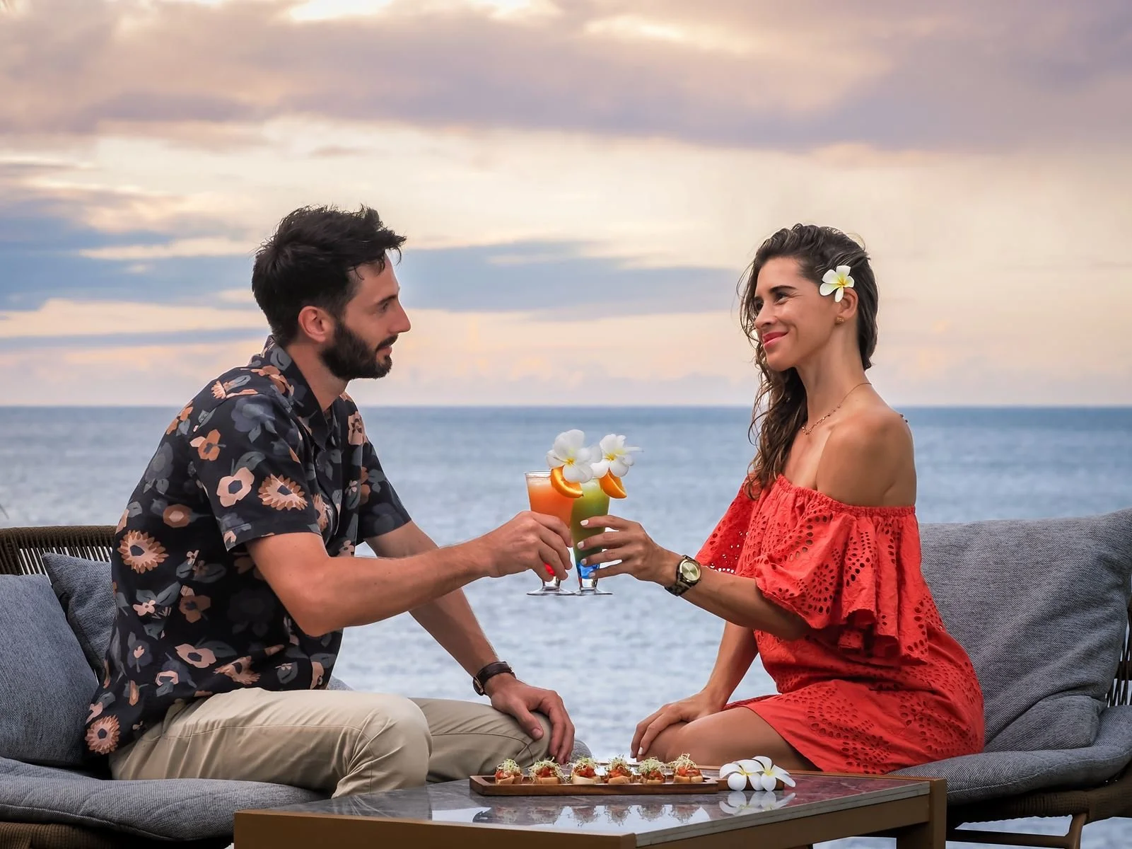 Un couple assis sur un canapé en extérieur par une mer, échangeant un toast avec des cocktails, en portant des vêtements d'été et des fleurs dans les cheveux, avec un plateau de petits snacks sur la table.