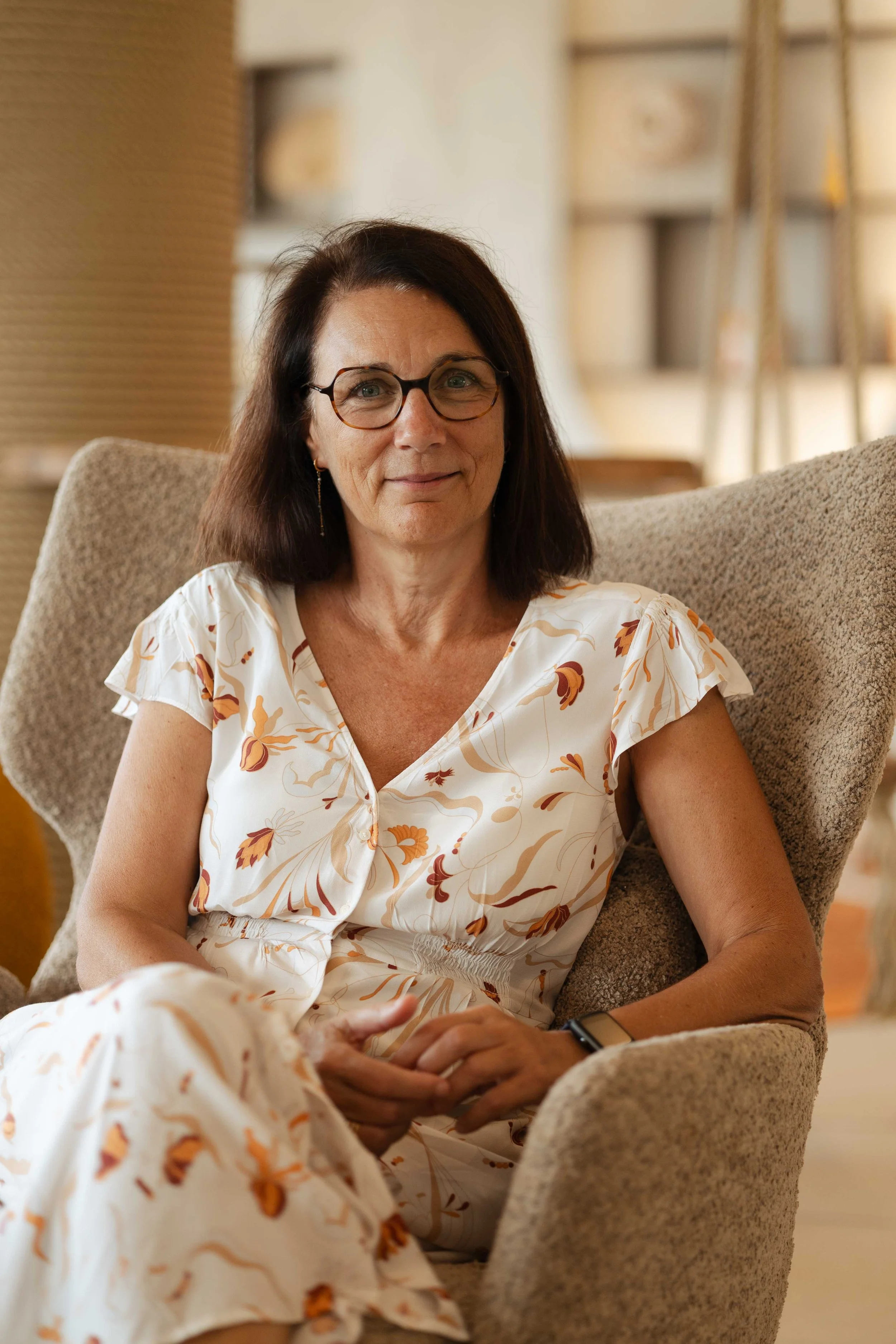 Une femme assise dans un fauteuil en tissu beige, portant une robe blanche à motifs floraux orange et beige, avec des lunettes noires et un bracelet noir, dans un intérieur chaleureusement éclairé.