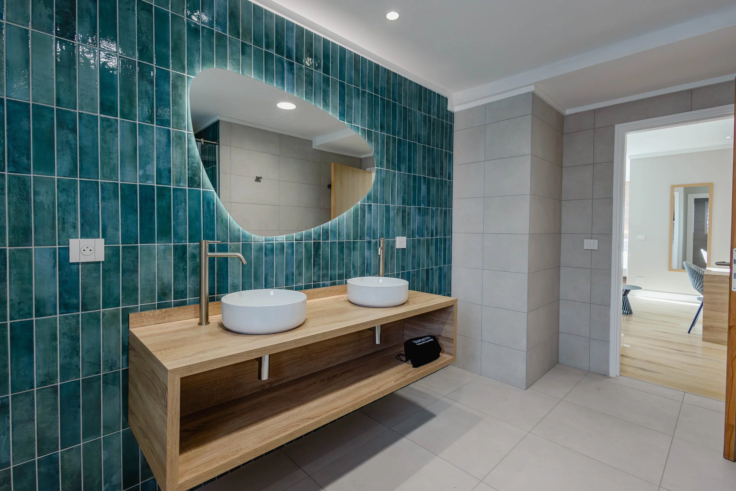Salle de bains moderne avec un grand miroir au-dessus d'un double lavabo en céramique blanc sur un plan de travail en bois avec une armoire ouverte en dessous. Le mur derrière le miroir est recouvert de carreaux turquoise. Il y a deux prises électriq