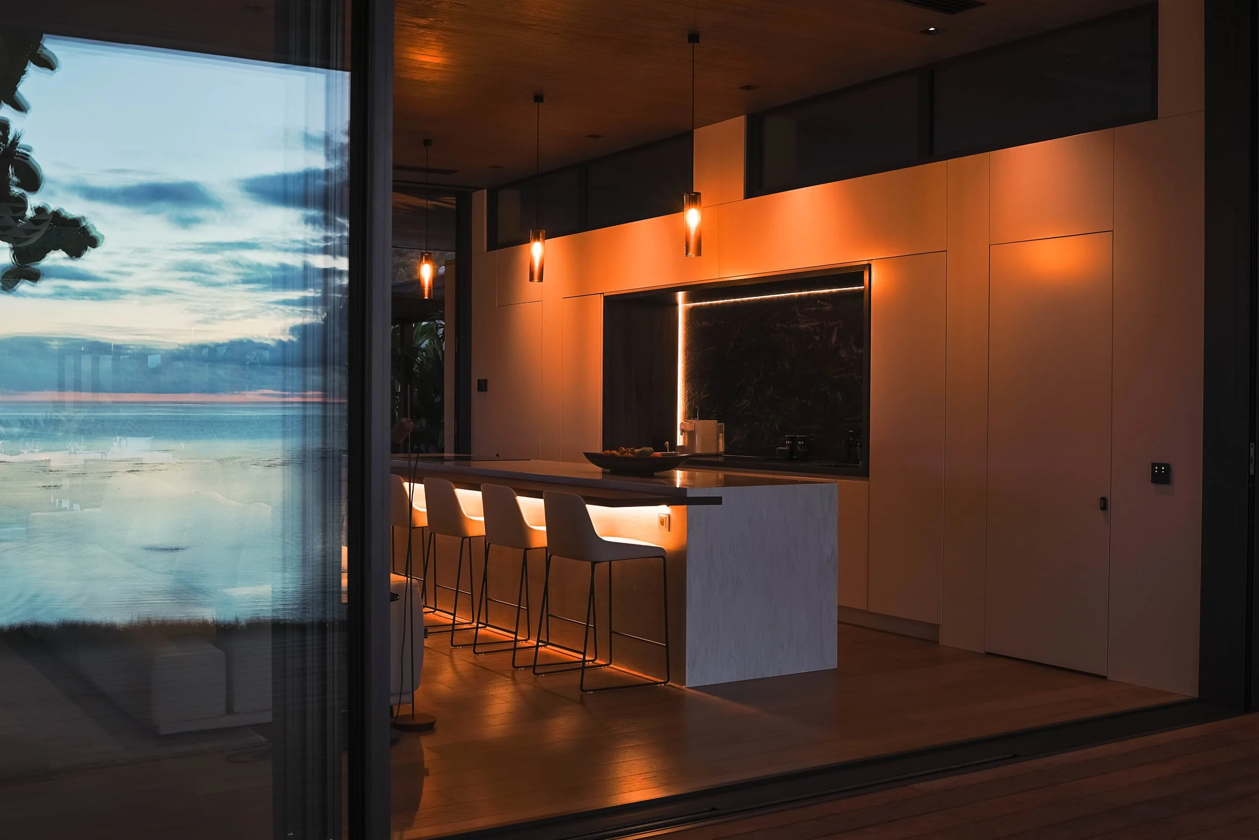 Intérieur moderne d'une cuisine avec îlot, chaises, éclairage chaleureux, vue sur la mer par une grande fenêtre ou porte en verre.
