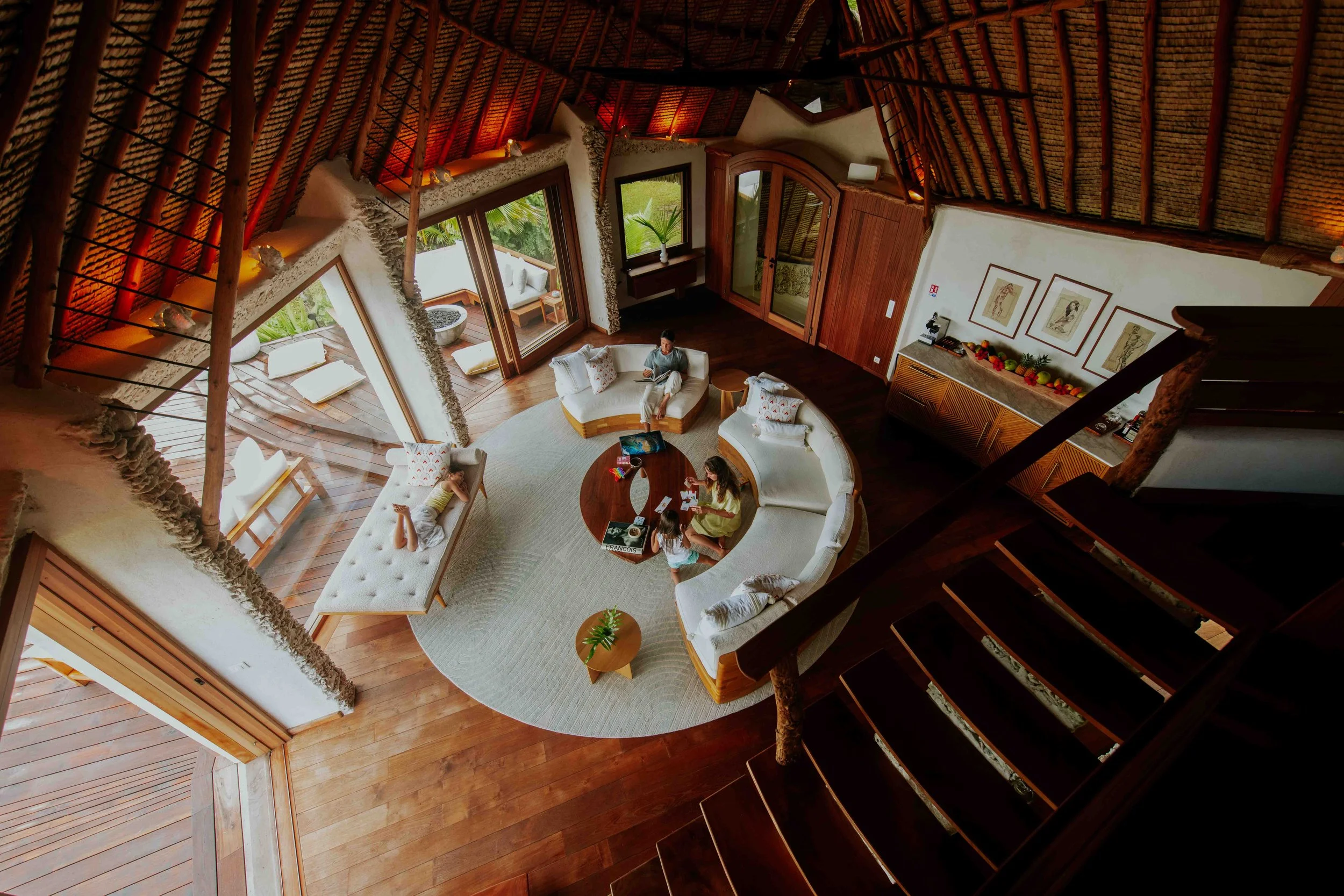 Vue intérieure d'un salon dans une maison de style tropical. Quatre personnes, probablement une famille, sont assises autour d'une table ronde en train de jouer ou de discuter, avec une grande banquette blanche et une autre personne sur un canapé. Le
