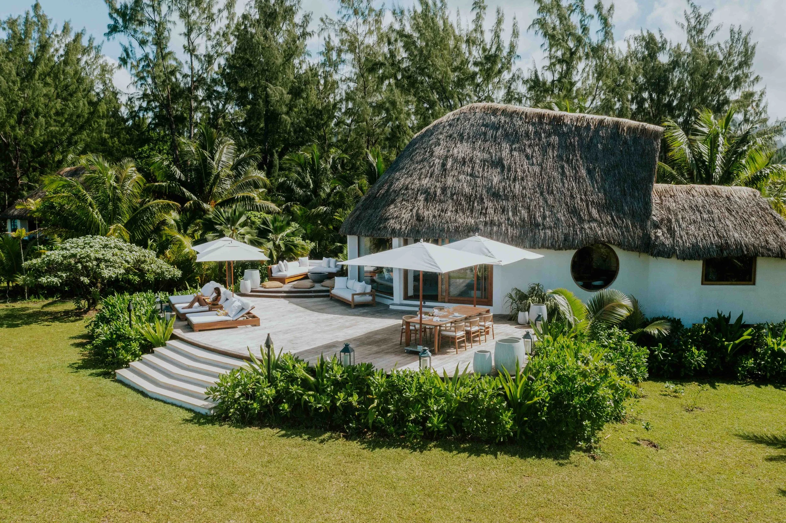 Une maison au toit de chaume avec une terrasse et des parasols blancs, entourée de végétation tropicale et un jardin verdoyant.