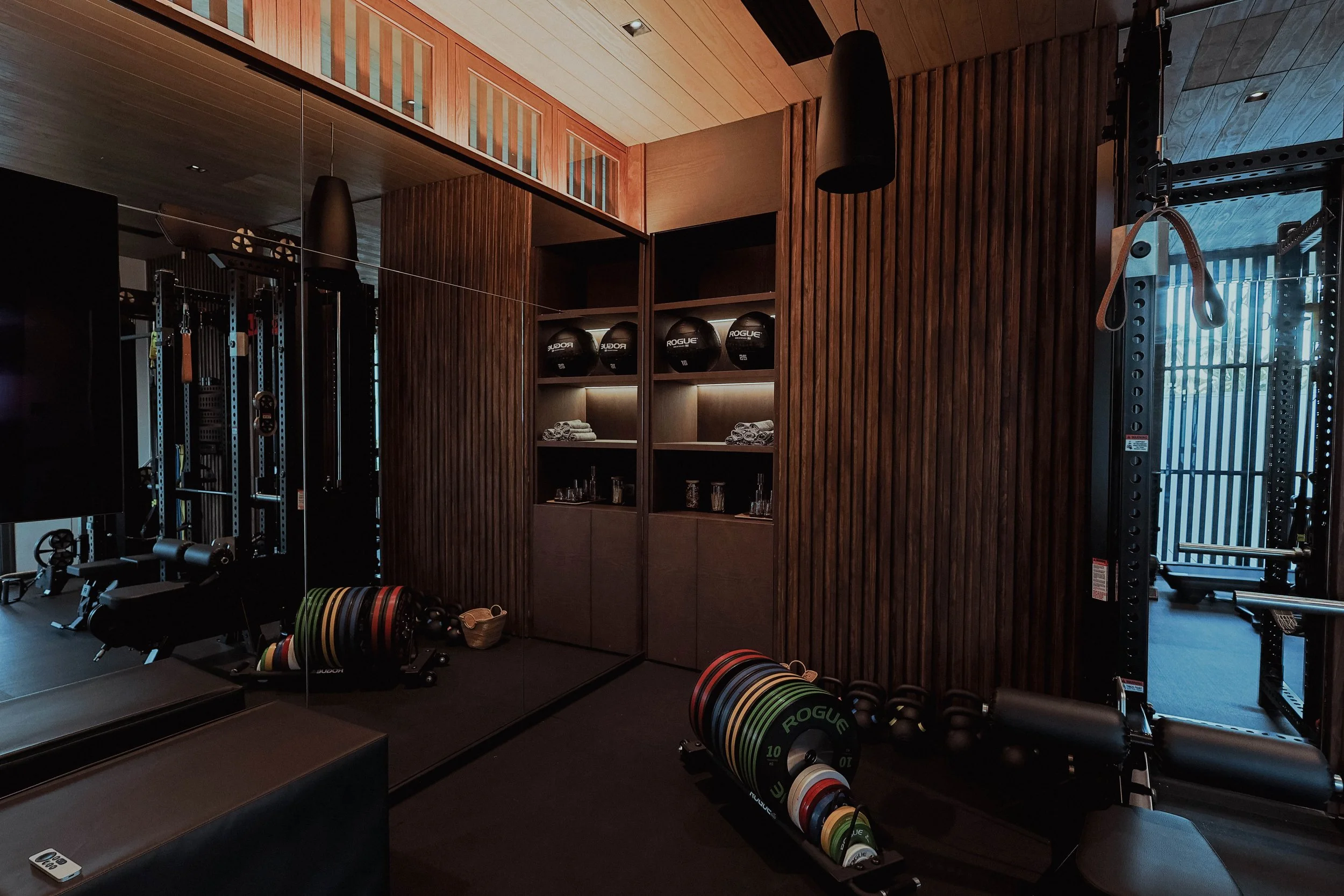 Salle de sport avec équipement de musculation, ballots de poids, kettlebells, et armoires avec des tapis et des balles de médecine, décor en bois.