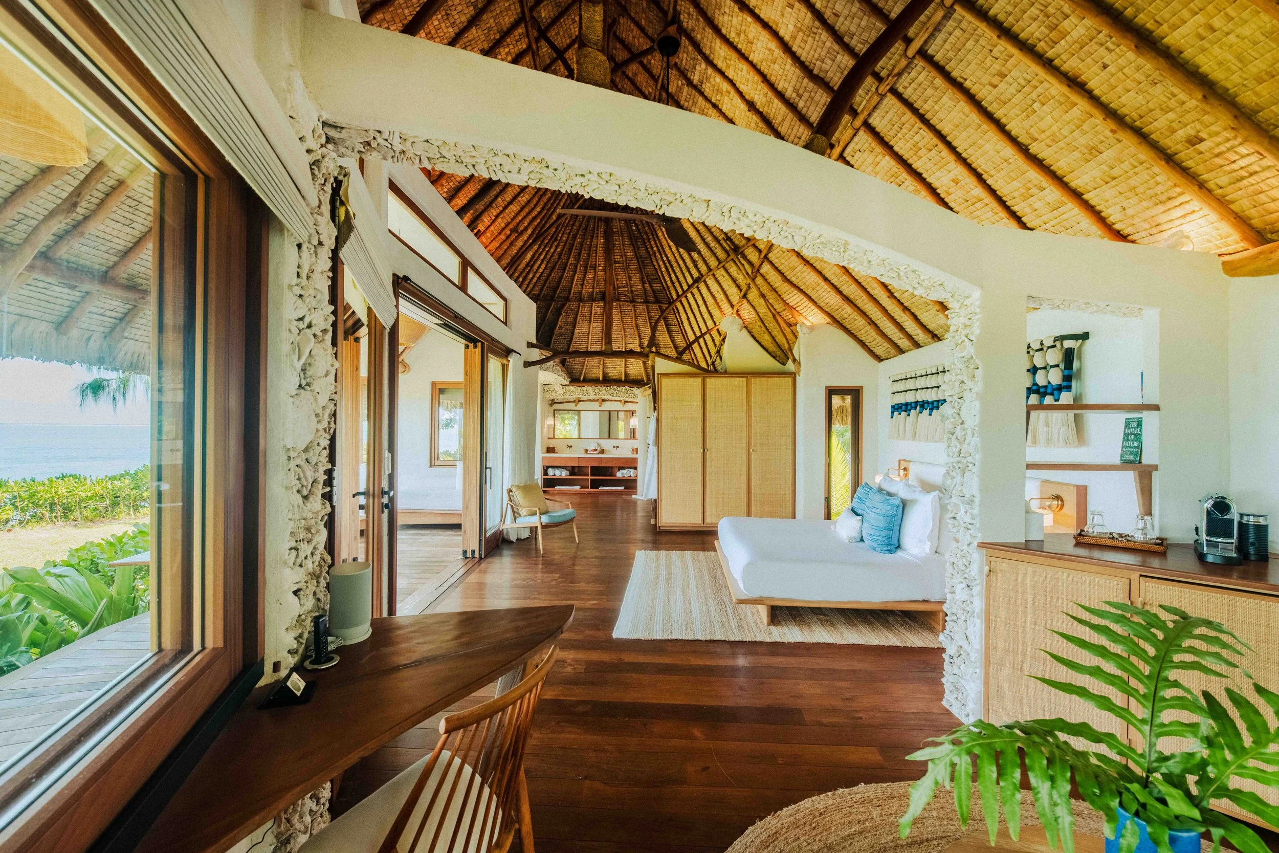 Intérieur d'une villa tropicale avec mur en pierre, plafond en bois de roseau, meubles en bois clair, canapé blanc avec coussins bleus, grand balcon avec vue sur la mer, plantes vertes, décor naturel et lumineux.
