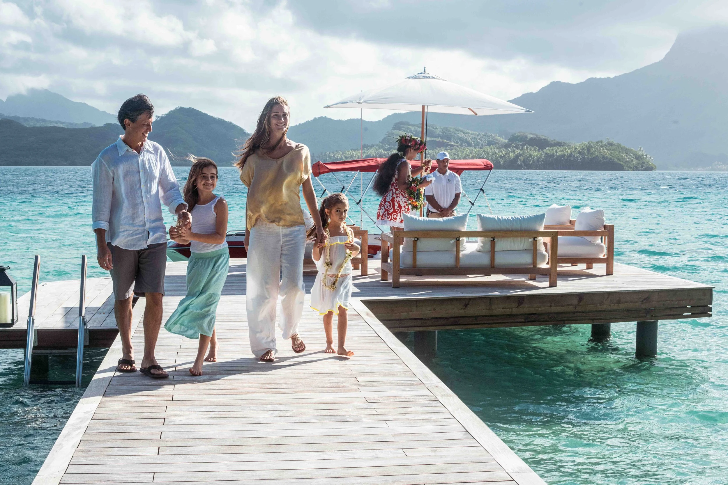 Une famille se promenant sur une jetée en bois au bord d'une mer turquoise, avec des montagnes en arrière-plan, sous un ciel partiellement nuageux.