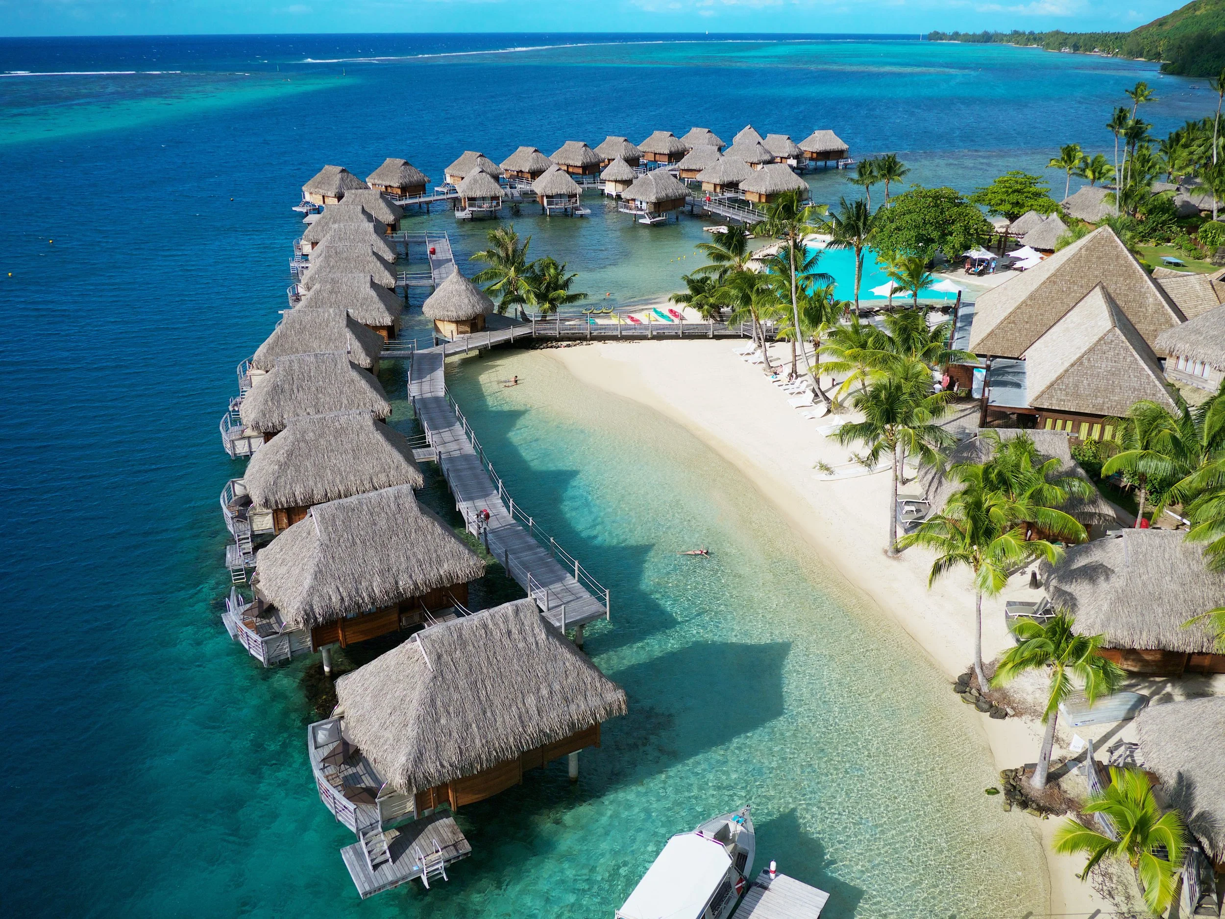 Vue aérienne d'un complexe hôtelier avec bungalows sur pilotis, plage de sable blanc, piscines, palmiers, et eaux turquoise.